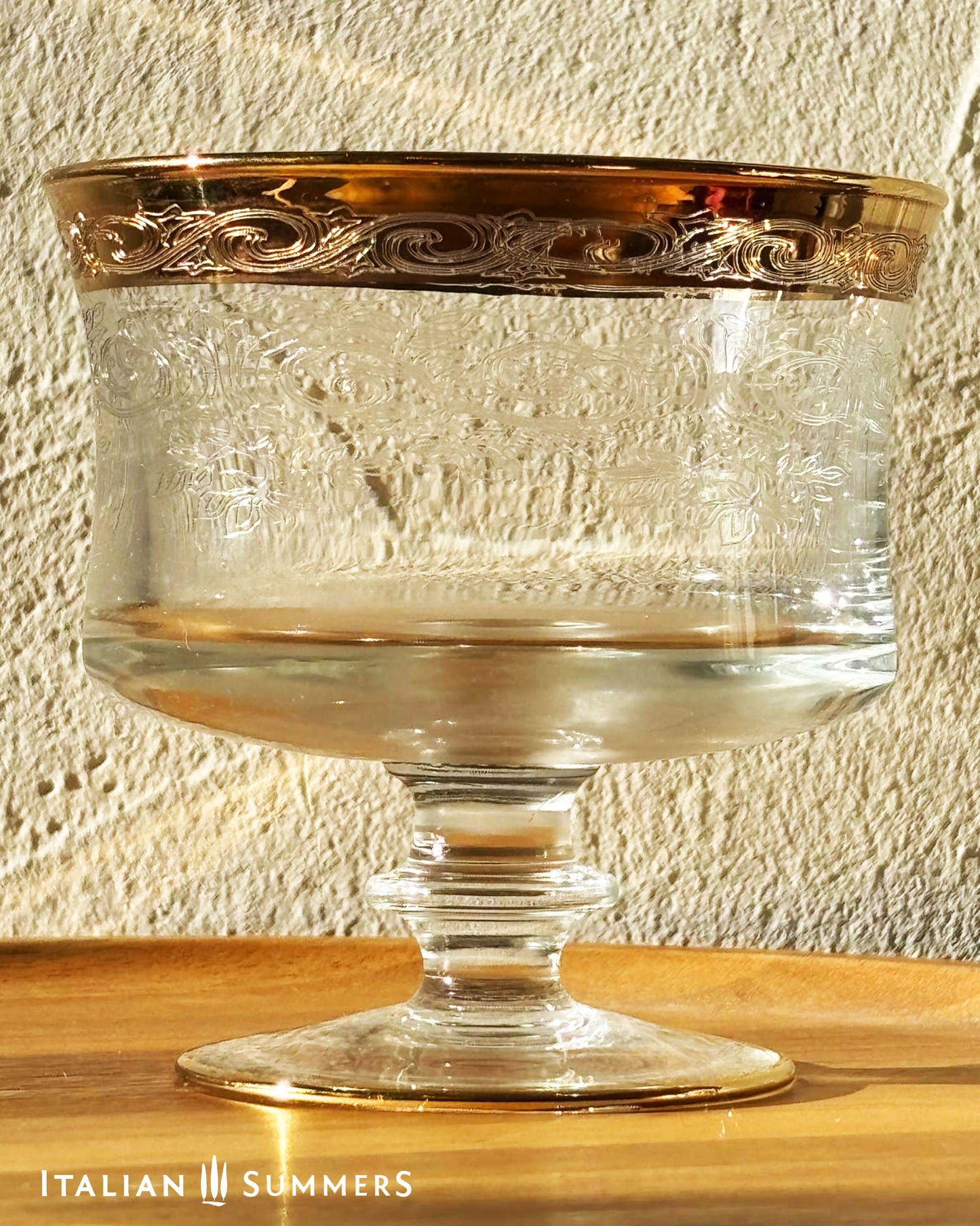  Glass coupe with gold rim and ornate floral filigree engravings on a wooden surface and textured wall background