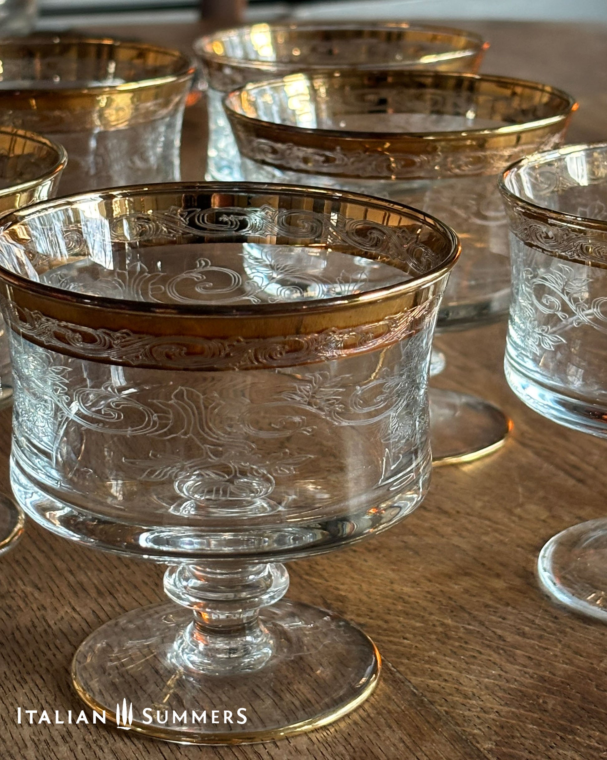Set of clear glass dessert coupes with gold rims and engraved floral filigree patterns  on a wooden surface, branded 'Italian Summers'.