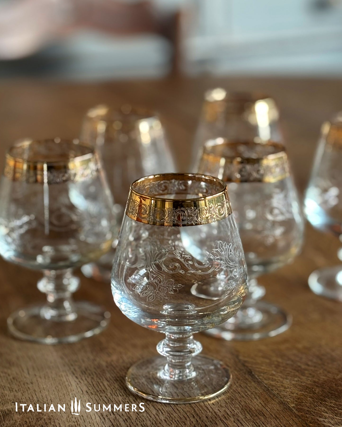 Set of clear glass goblets with gold rims on a wooden surface, branded 'Italian Summers'.