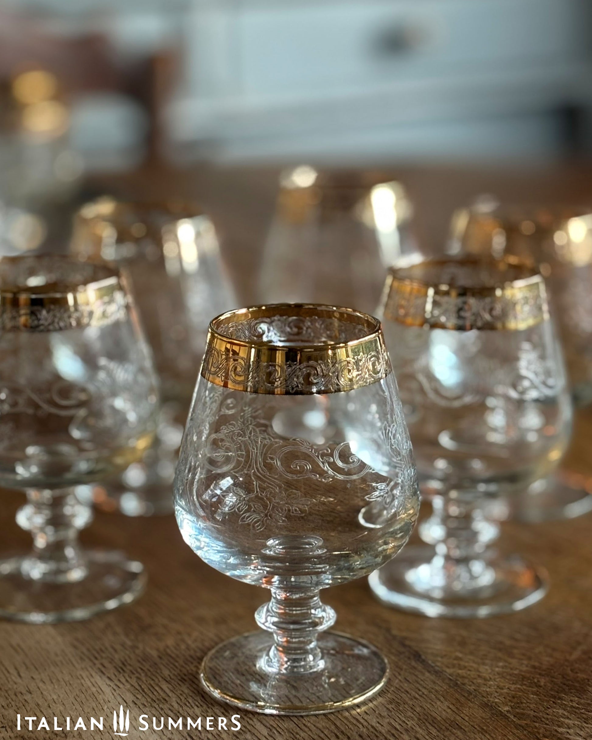 Set of clear glass goblets with gold rims on a wooden surface, branded 'Italian Summers'.