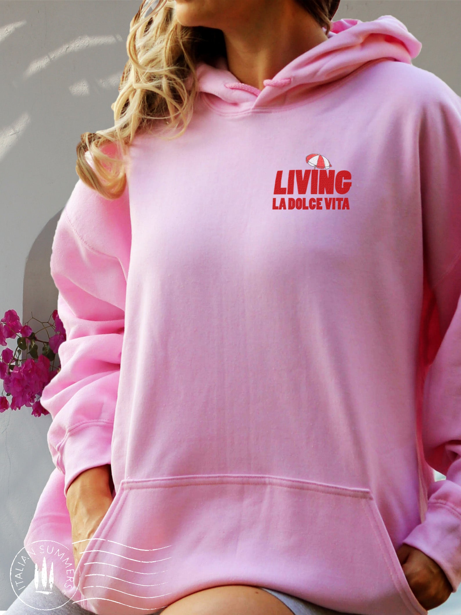 A n Italy-inspired, soft and comfortable made to order  Hoodie . Pink cotton blend with an embroidered red decoration text  LIVING LA DOLCE VITA with a red and white beach umbrella on top.  Large print of the same text on the back. Front pocket