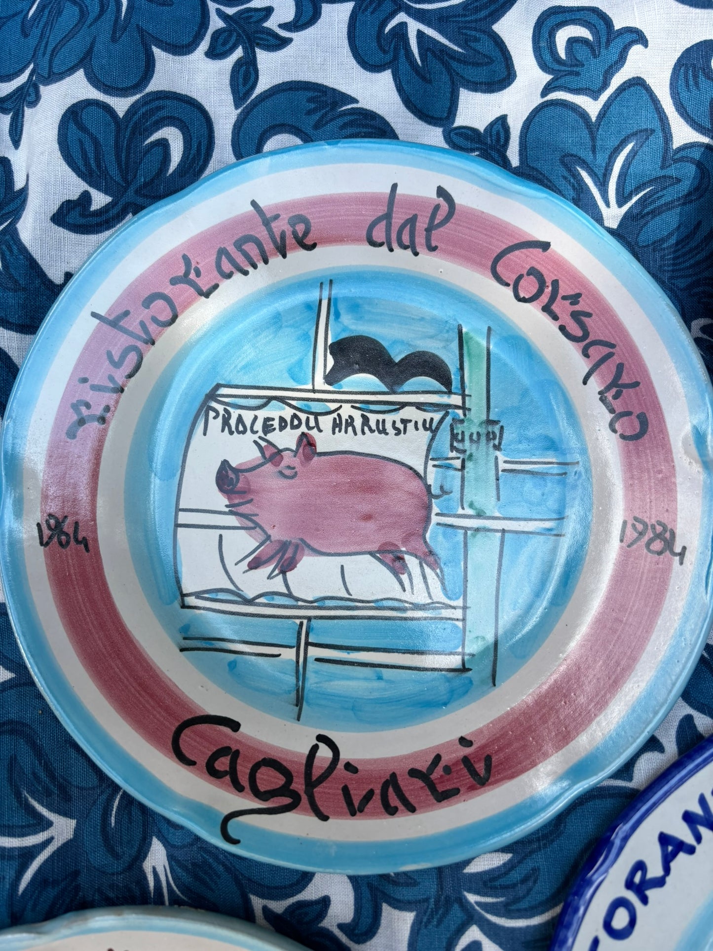 Italian ceramic plate with a handpainted pig illustration and text Ristorante dal Corsaro Cagliari on a blue and white patterned tablecloth