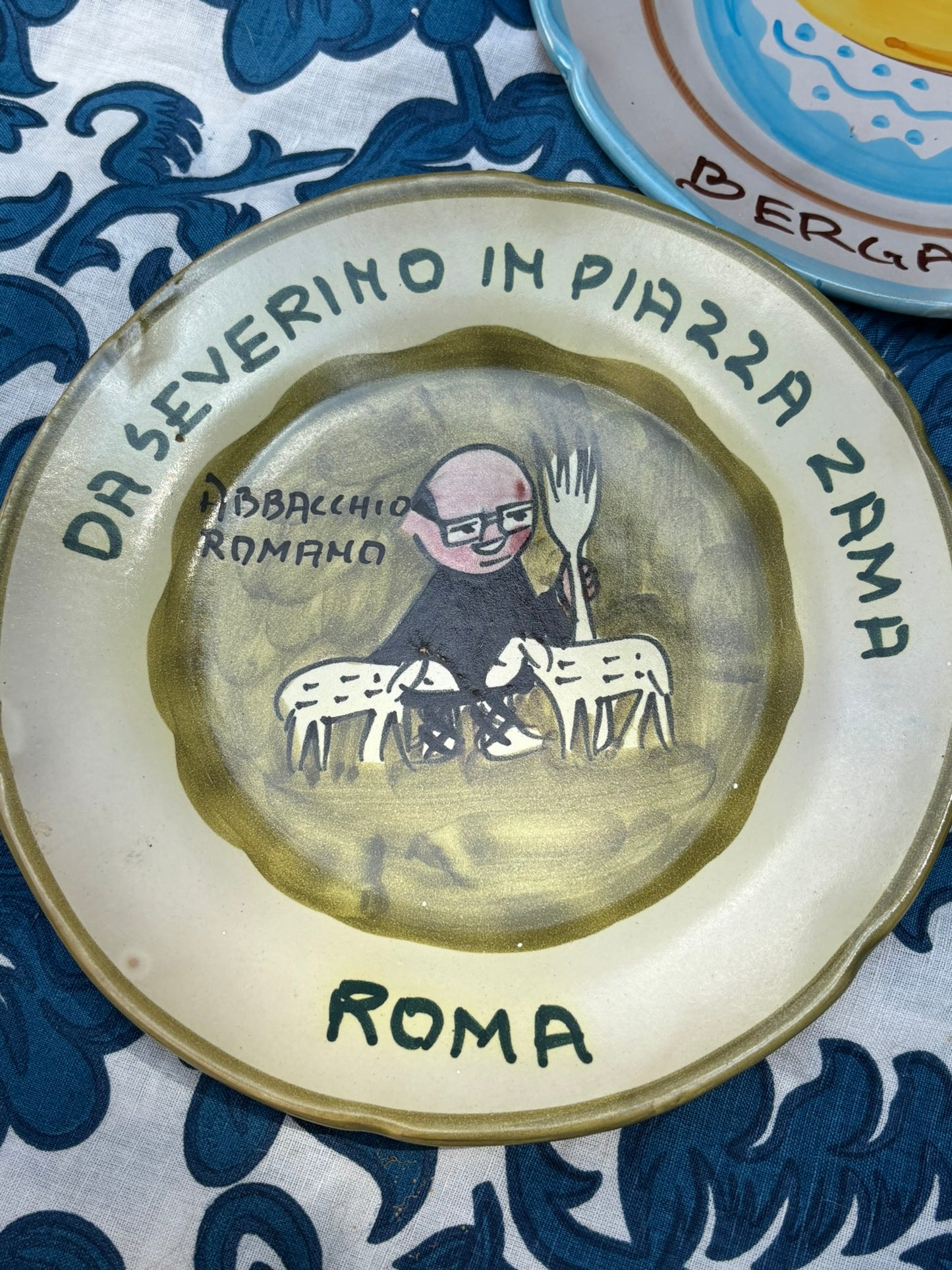 Italian vintage ceramic plate with a handpainted cartoonish character and Italian text on a blue and white patterned tablecloth