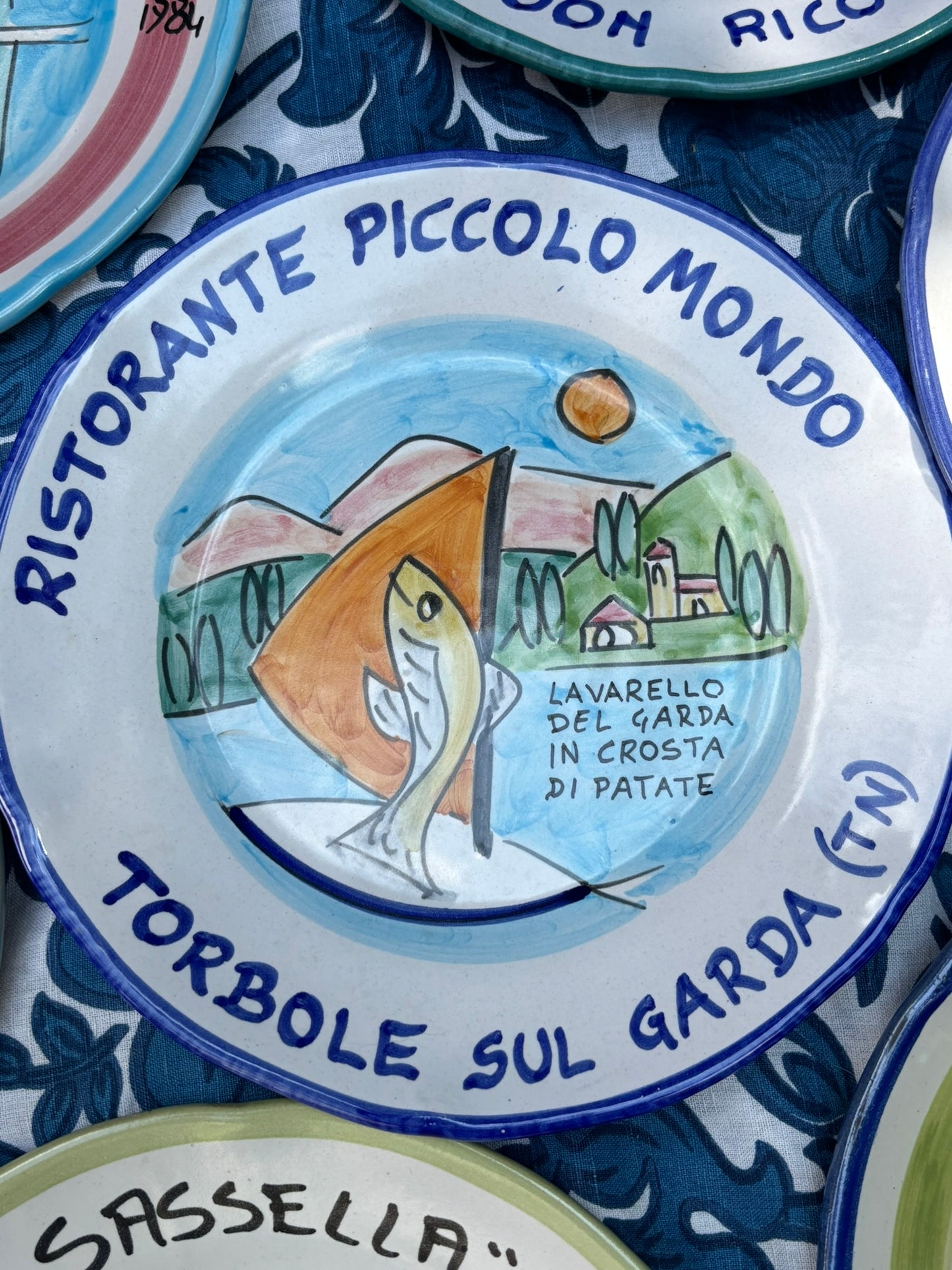 Italian Ceramic plate with handpainted  decorative design and Italian text Ristorante Piccolo Mondo Torbole sul Garda, featuring a fish and Italian landscape, surrounded by other similar plates.
