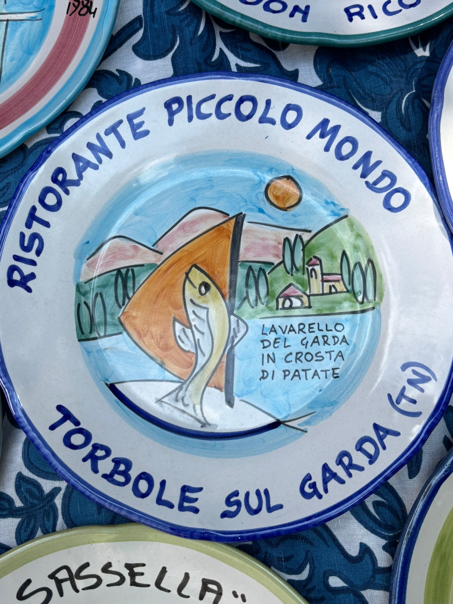 Italian Ceramic plate with handpainted  decorative design and Italian text Ristorante Piccolo Mondo Torbole sul Garda, featuring a fish and Italian landscape, surrounded by other similar plates.