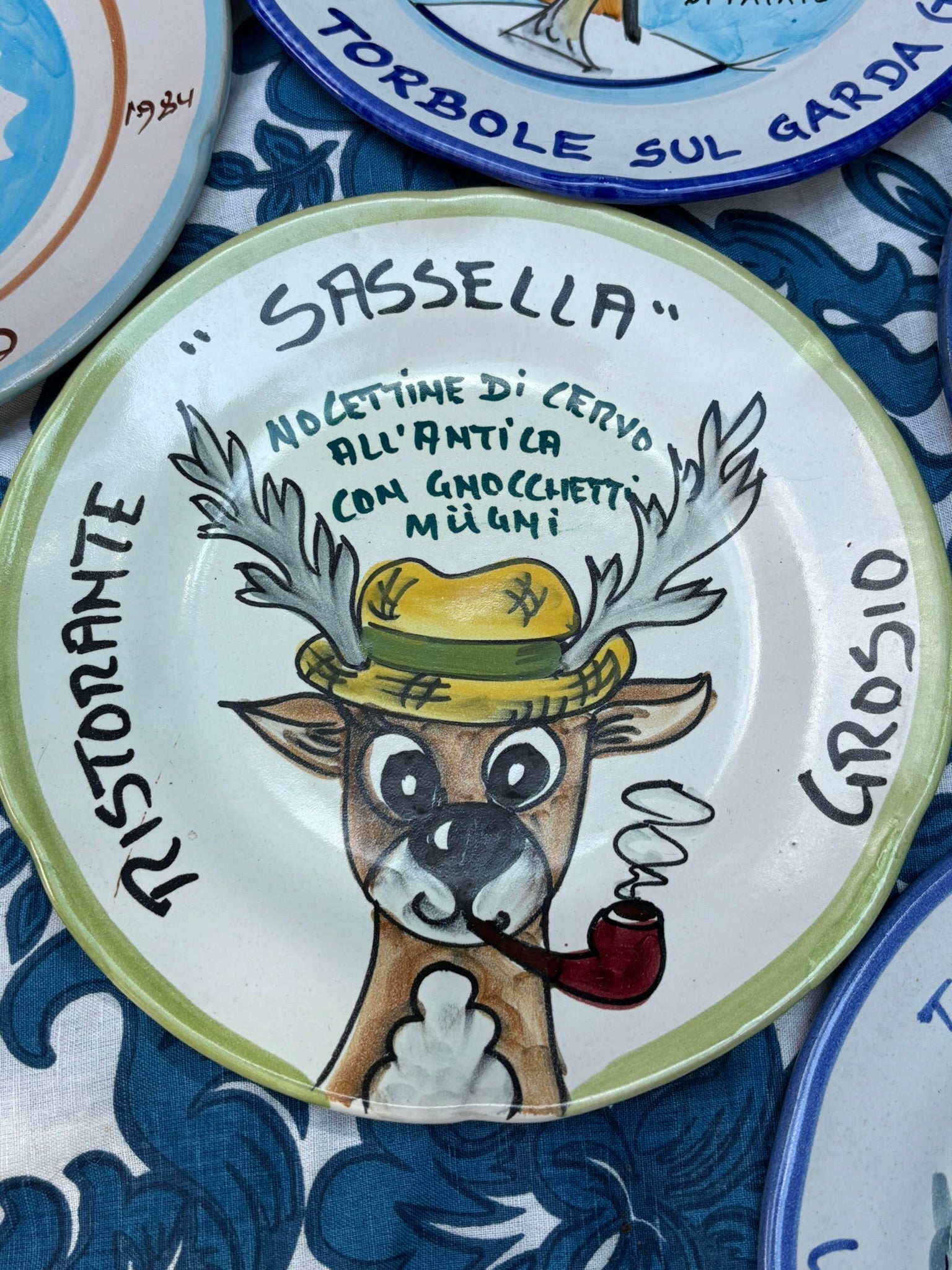 Italian ceramic plate with a handpainted cartoon deer wearing a hat and smoking a pipe, and Italian text Ristorante Sassella Grosio and an Italian recipe, surrounded by other plates.