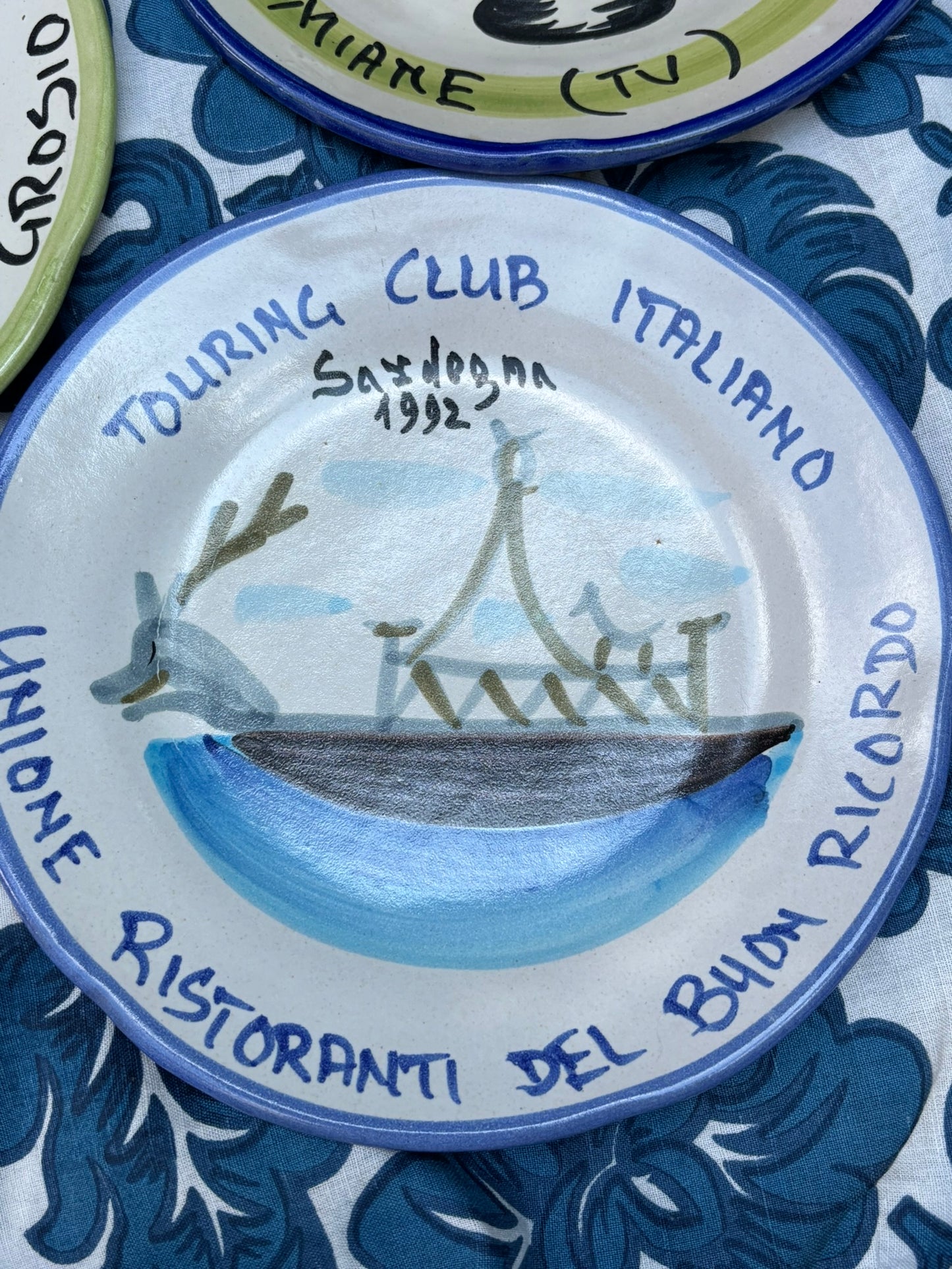 Italian ceramic plate with 'Touring Club Italiano Sardegna' text and design of a boat in the sea on a blue and white patterned tablecloth