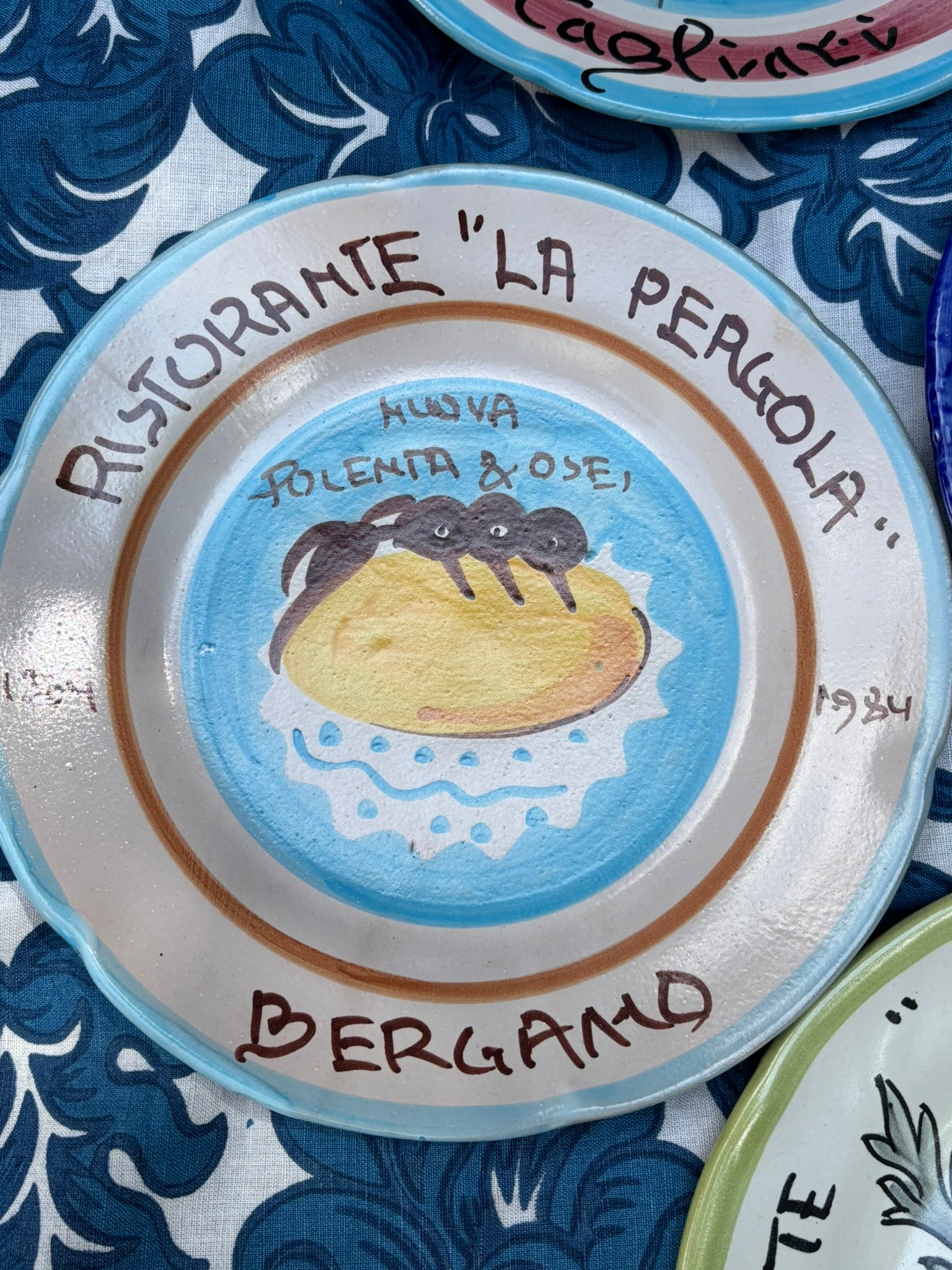 Italian vintage ceramic plate with 'Ristorante La Pergola' text and a handpainted food illustration on a blue and white patterned  talecloth.