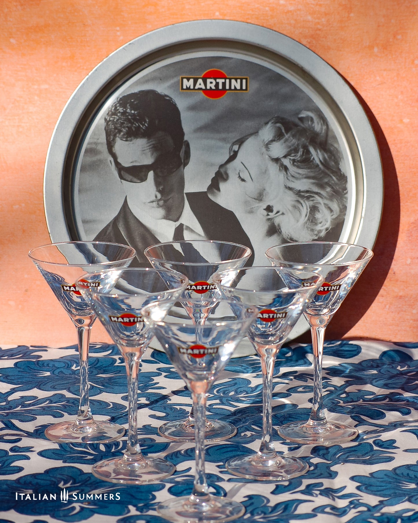 Set of Martini glasses on a table with a tray featuring Charlize Theron and Max Parrish advert for Martini in the background.