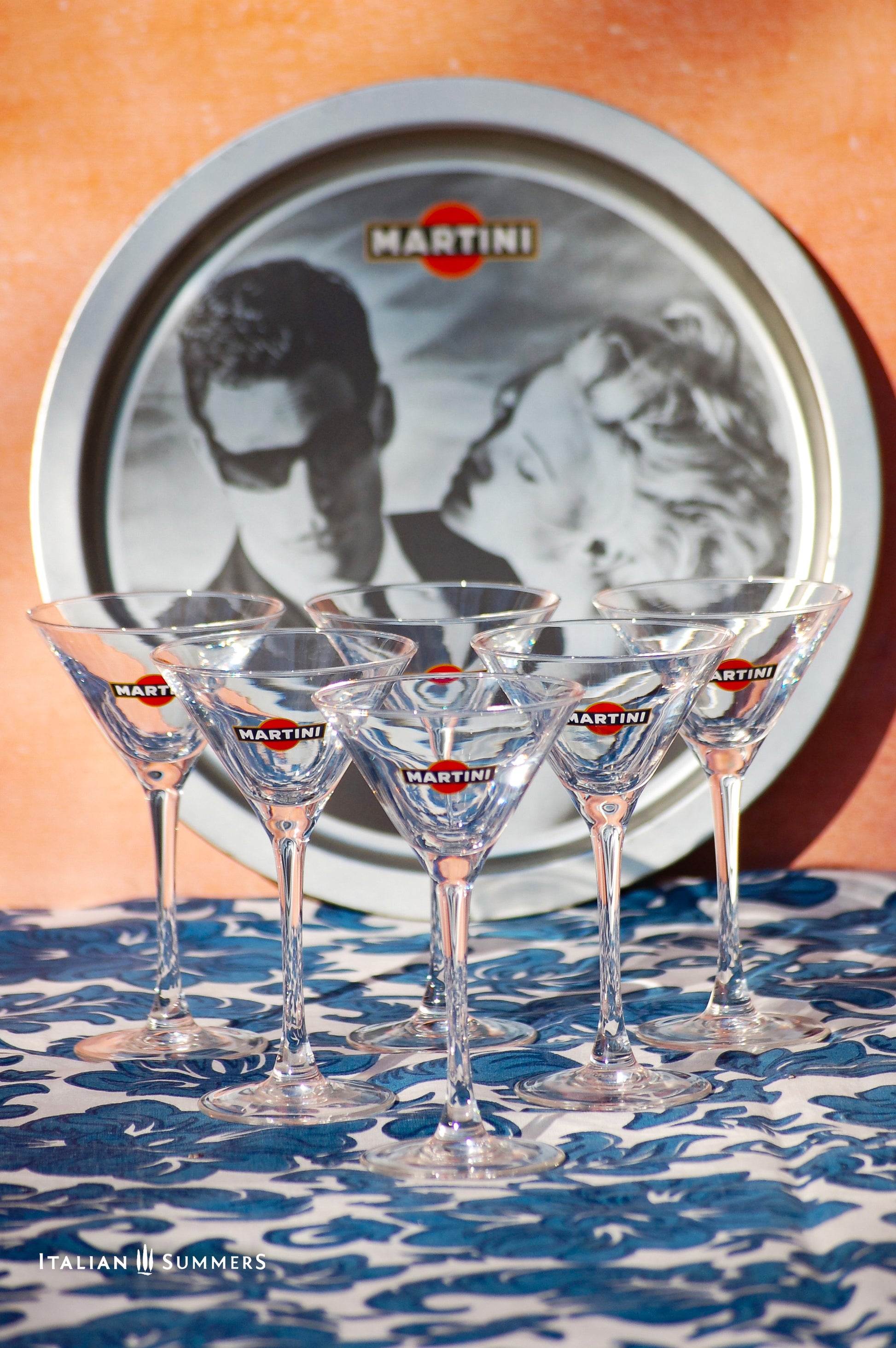 Set of Martini glasses on a table with a tray featuring Charlize Theron and Max Parrish advert for Martini in the background.