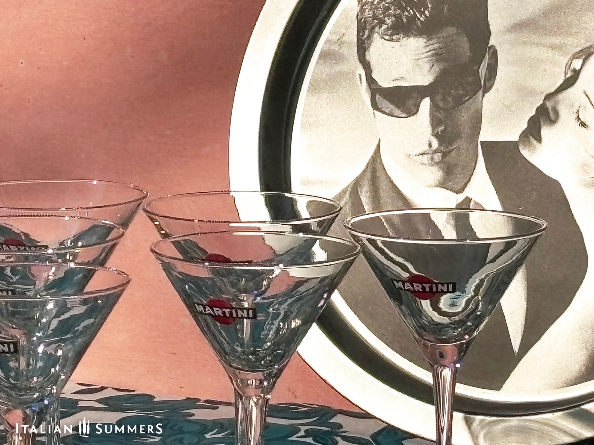 Set of Martini glasses on a table with a tray featuring Charlize Theron and Max Parrish advert for Martini in the background.