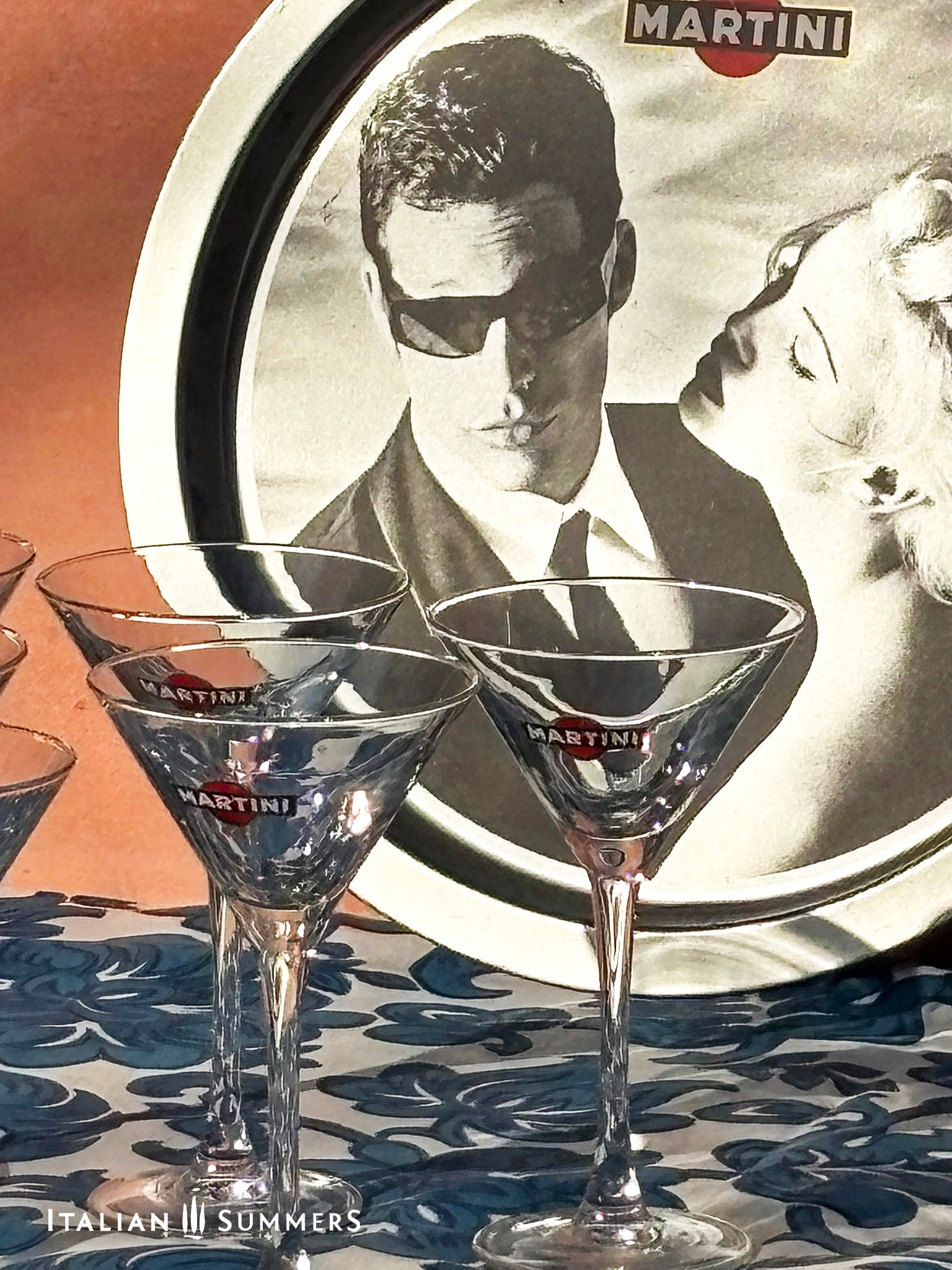 Set of Martini glasses on a table with a tray featuring Charlize Theron and Max Parrish advert for Martini in the background.