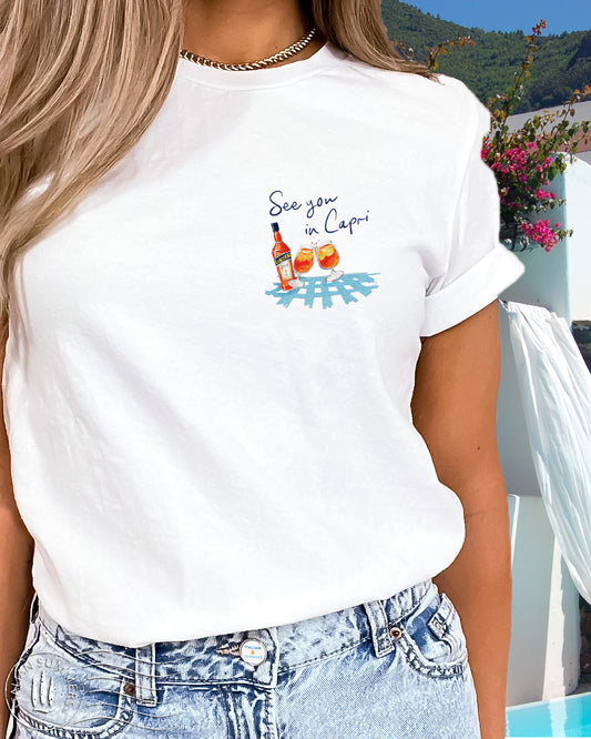 White cotton T shirt with an embroidered illustration on the left chest of two Aperol Spritz glasses in red-orange colors and a Aperol bottle on a blue checkered tablecloth. Above, a Navy blue embroidered quote: See you in CAPRI