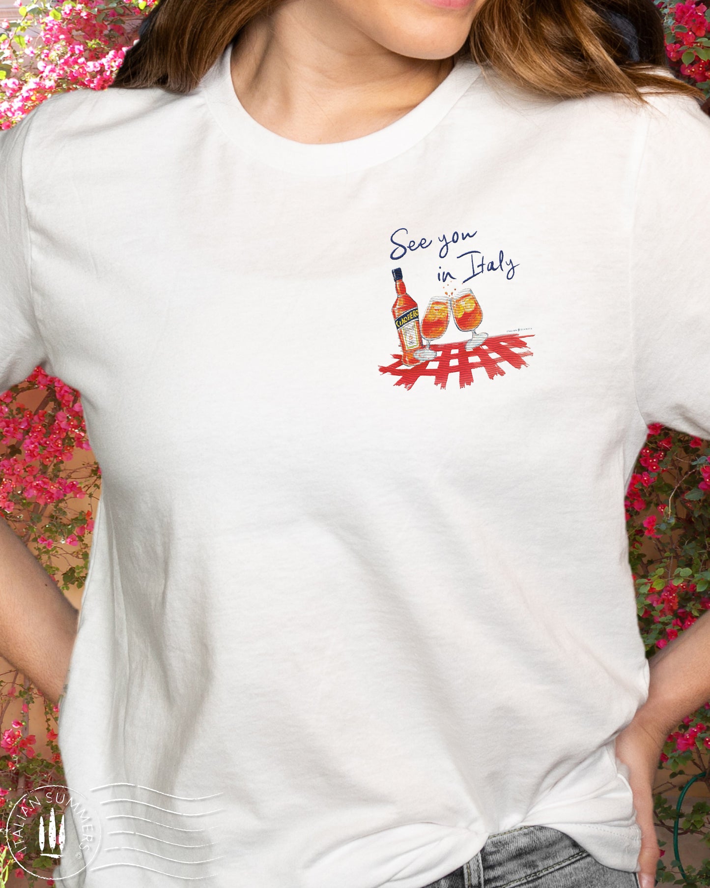 White cotton T shirt with an embroidered illustration on the left chest of two Aperol Spritz glasses in red-orange colors and a Aperol bottle on a red checkered tablecloth. Above, a Navy blue embroidered quote: See you in Italy Desgned and sold by Italian Summers
