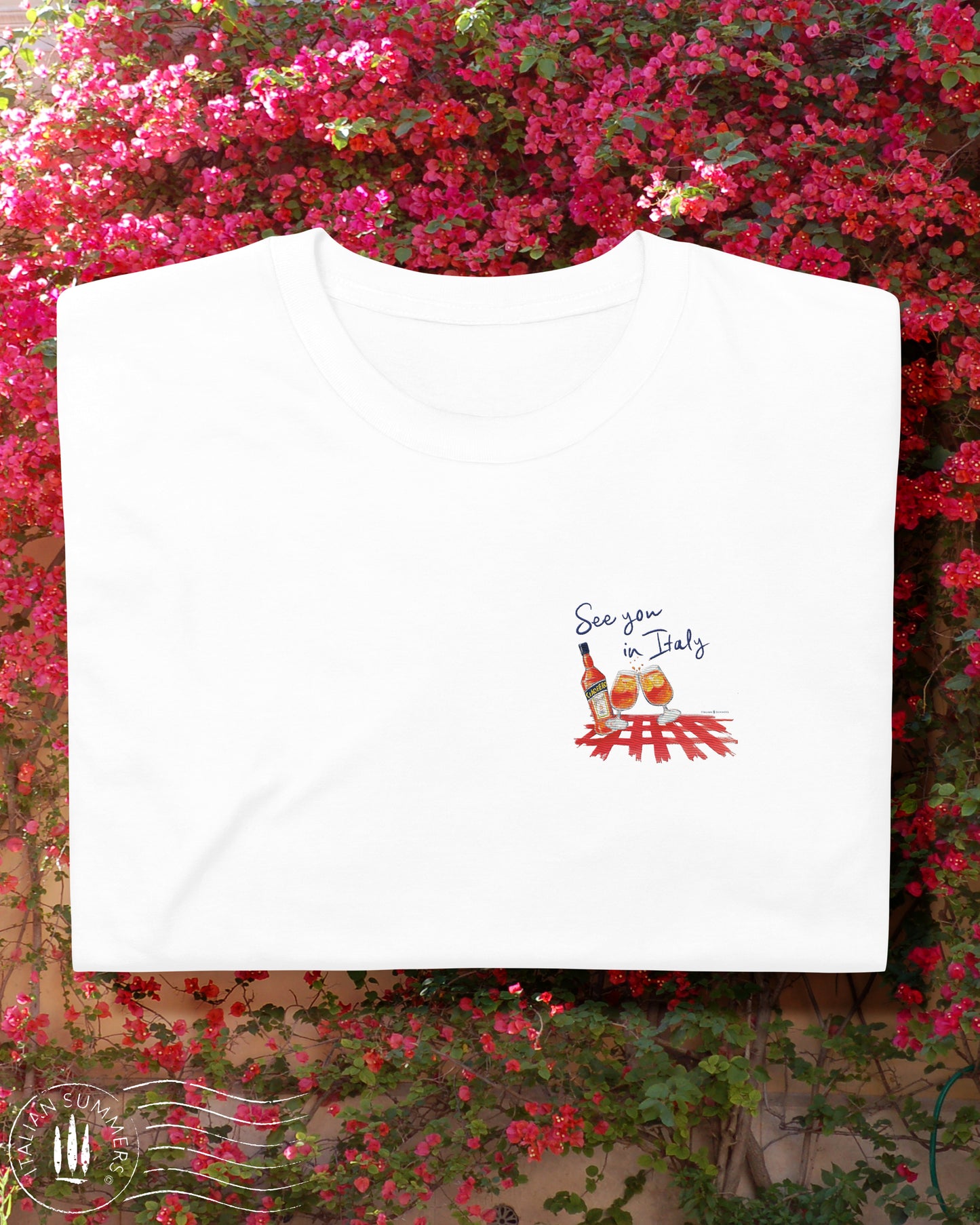 White cotton T shirt with an embroidered illustration on the left chest of two Aperol Spritz glasses in red-orange colors and a Aperol bottle on a red checkered tablecloth. Above, a Navy blue embroidered quote: See you in Italy Desgned and sold by Italian Summers