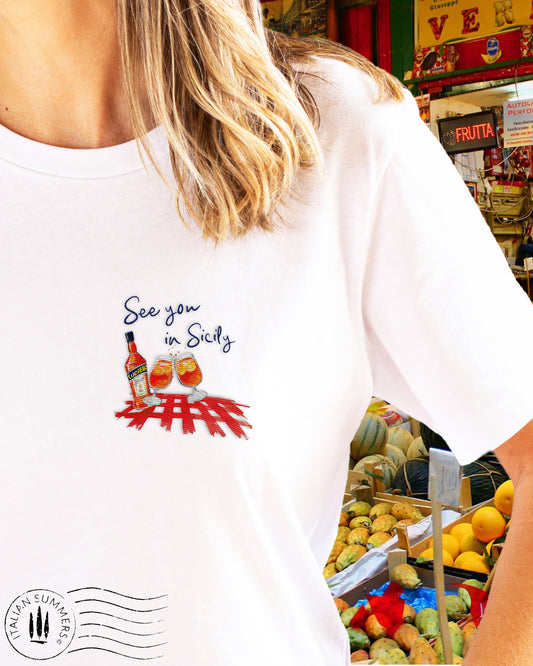 A white crew-neck cotton T Shirt with an emroidered decoration on the left chest, depicting a pair of Aperol Spritz glasses, a Bottle of Aperol  over a red checkered tablecloth pattern. The quote "See you in Sicily" is above the decoration.