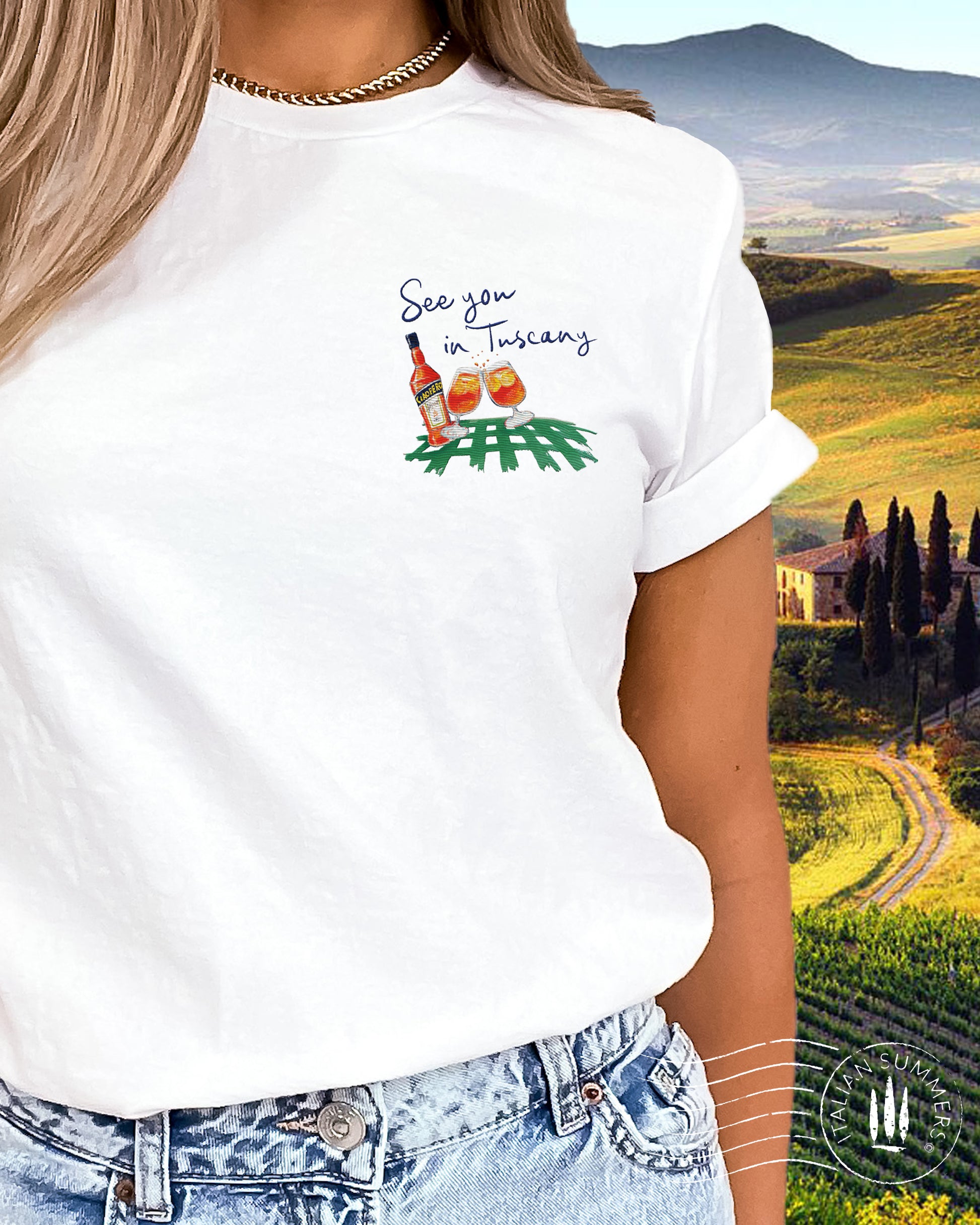 A white crew-neck cotton T Shirt with an embroidered decoration on the left chest, depicting a pair of Aperol Spritz glasses, a Bottle of Aperol over a red or green checkered tablecloth pattern. The quote "See you in Tuscany" is above the decoration.