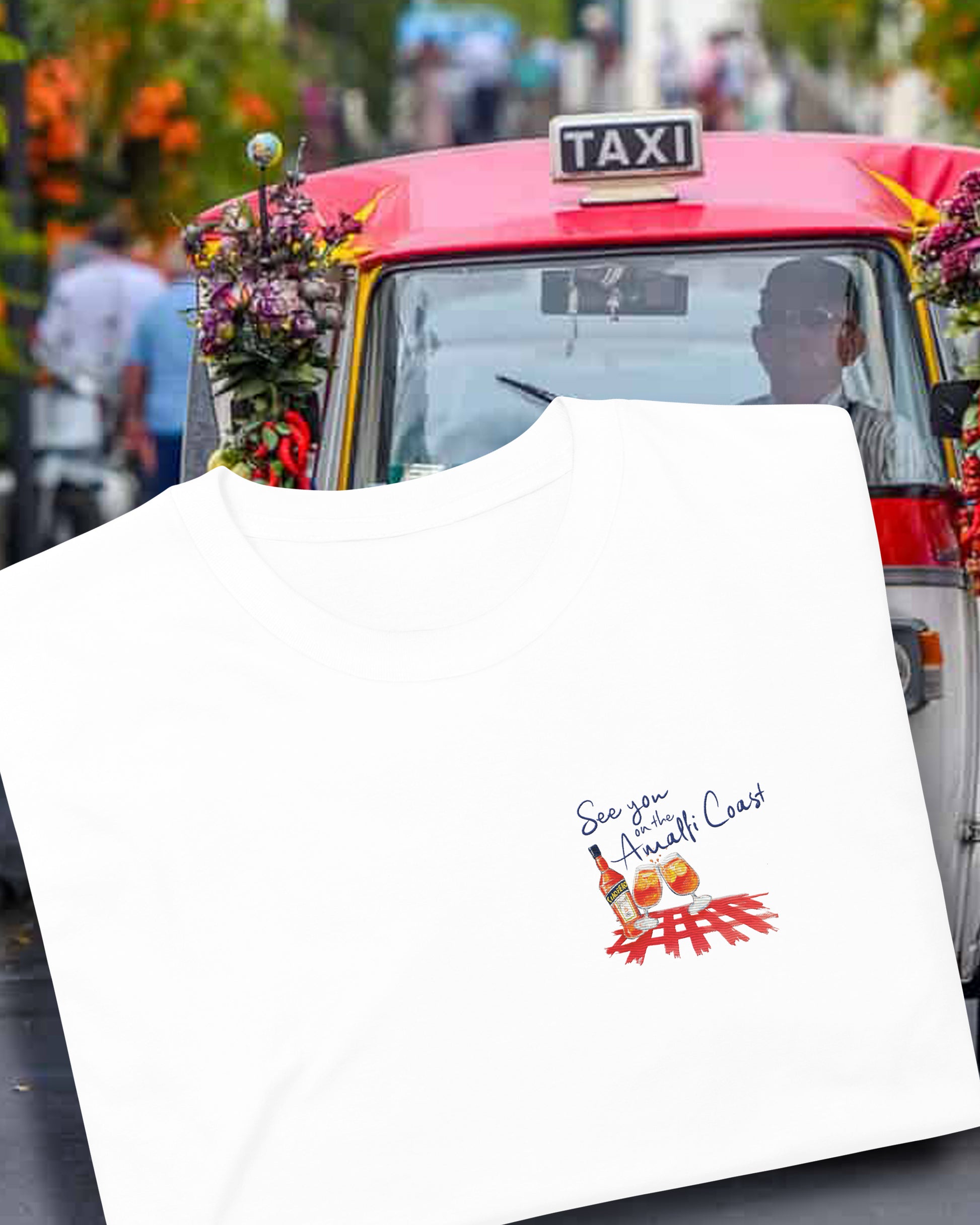 See you on the Amaaldi Coast. White cotton T shirt with an embroidered illustration on the left chest of two Aperol Spritz glasses in red-orange colors and a Aperol bottle on a red checkered tablecloth. Above, a Navy blue embroidered quote: See you on the Amalfi Coast.