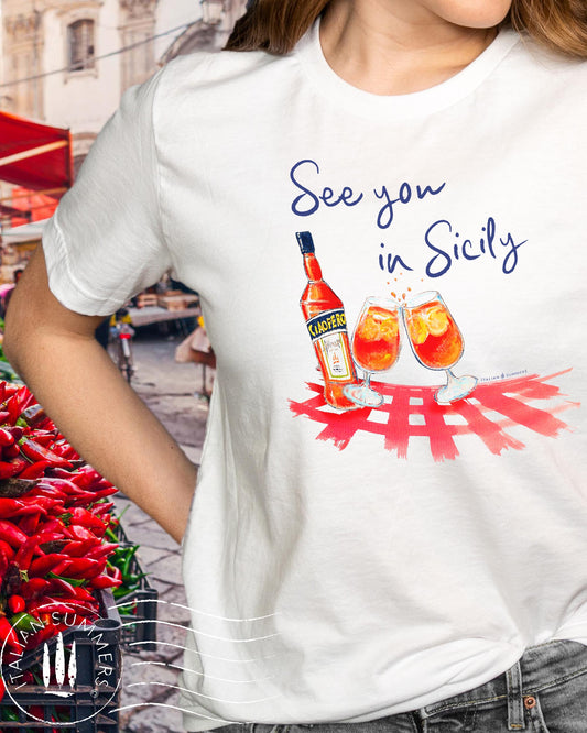 A white crew-neck cotton T Shirt with an printed decoration on the chest, depicting a pair of Aperol Spritz glasses, a Bottle of Aperol over a red or green checkered tablecloth pattern. The quote "See you in Sicily" is above the decoration.