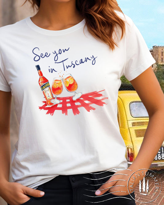 A white crew-neck cotton T Shirt with a printed decoration on the chest, depicting a pair of Aperol Spritz glasses, a Bottle of Aperol over a red checkered tablecloth pattern. The quote "See you in Tuscany" is above the decoration.