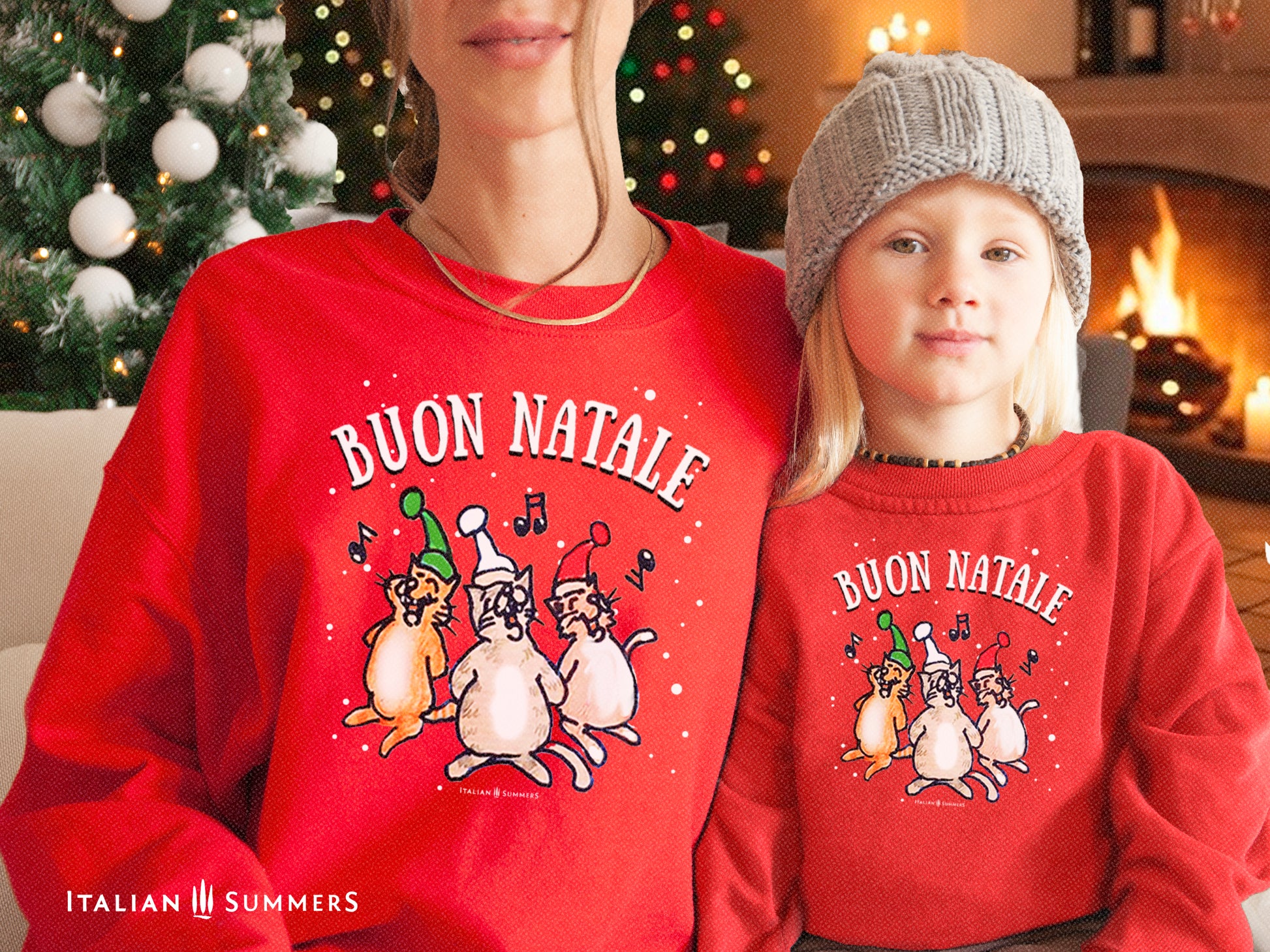 A mother and young blonde daughter both sit in a quaint room with a Christmas tree and lights and a fireplace, they both are wearing a red Christmas sweatshirt with a print of a drawing of three singing Italian cats in the snow with Christmas hats. Above the text: BUON NATALE.