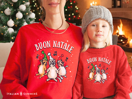 A mother and young blonde daughter both sit in a quaint room with a Christmas tree and lights and a fireplace, they both are wearing a red Christmas sweatshirt with a print of a drawing of three singing Italian cats in the snow with Christmas hats. Above the text: BUON NATALE.
