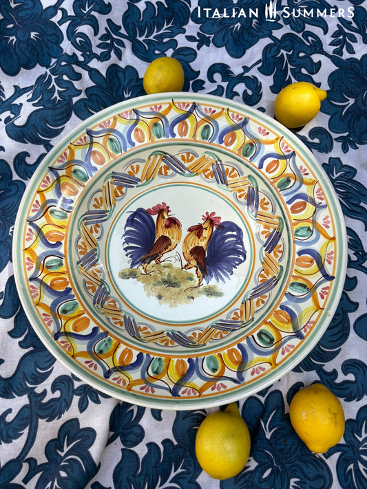 A large Apulian serving or decorative ceramic plate with rooster design on a blue patterned tablecloth with lemons.