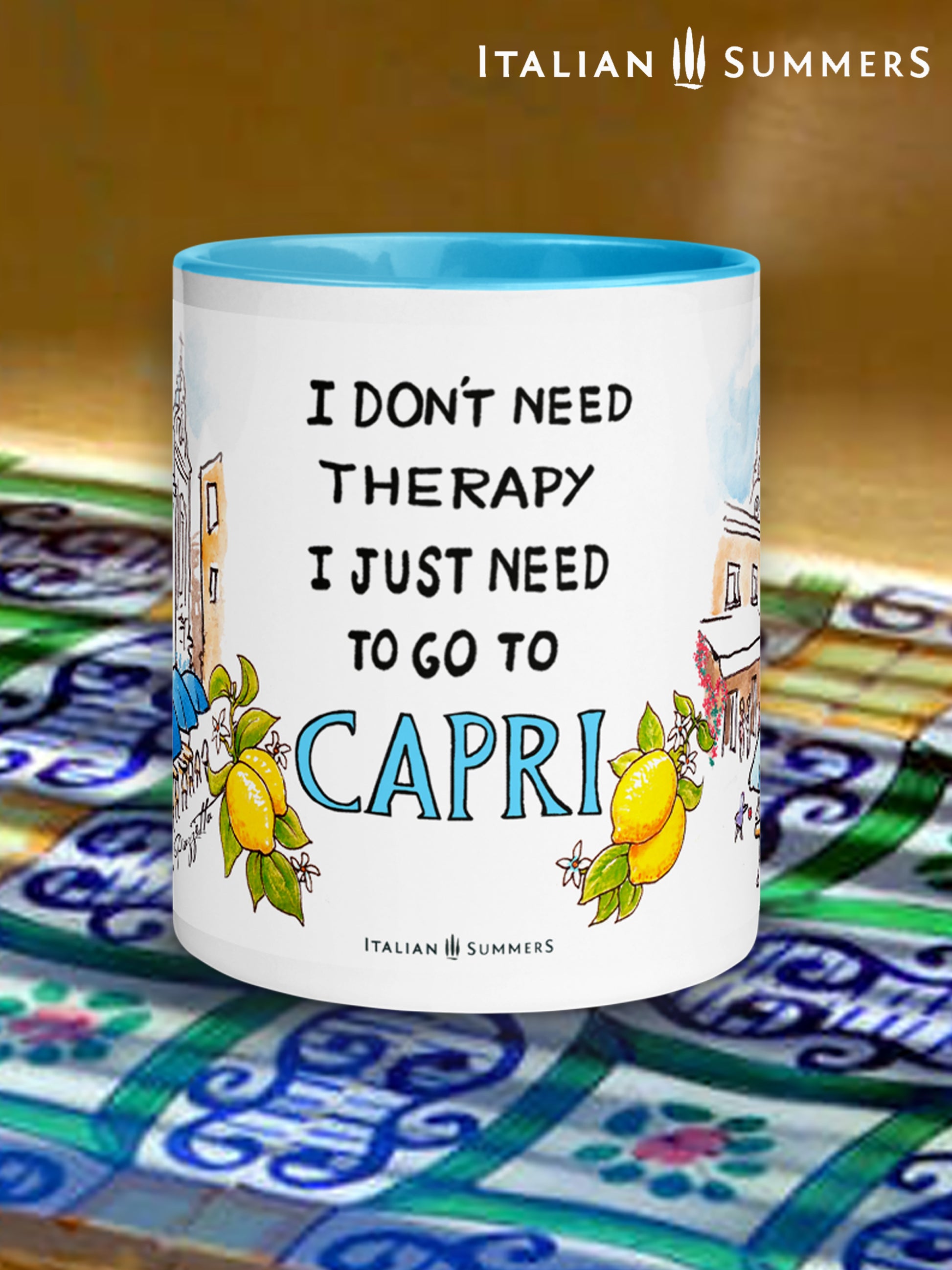 Italy-inspired white ceramic mug with blue colored handle and interior. Printed with a drawing of Capri's Piazzetta  with blue cafe Umbrellas and a crowd of shoppers strolling. The quote: I don't need therapy I just need to go to Capri is present . The word Italy is in blue Capital letters and flanked by bunches of Sorrento lemons with flowers. Made by Italian Summers