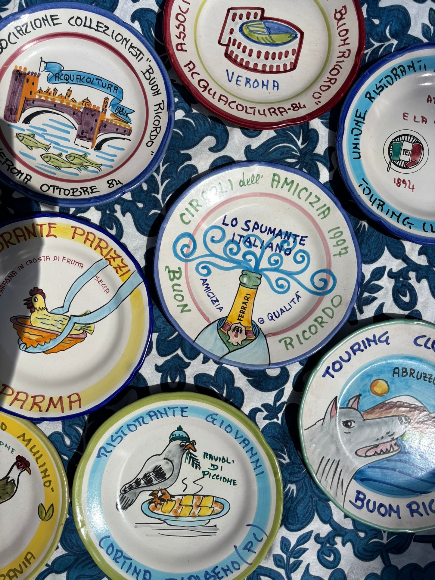 Collection of decorative Italian Buon Ricordo plates with various designs and text on a blue and white patterned background. 