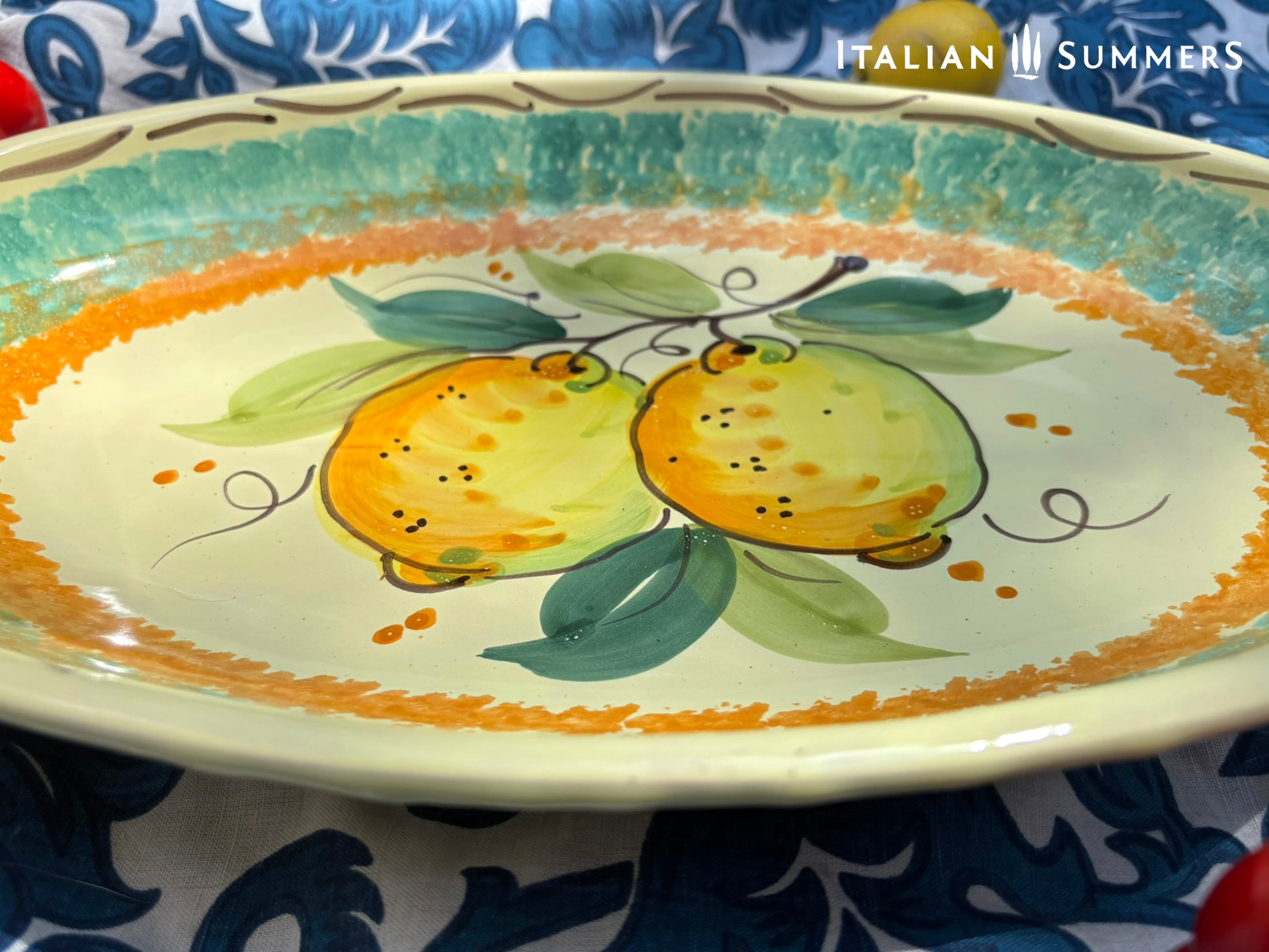 A large oval serving or Decorative Sicilian ceramic plate with alemon design on a blue floral tablecloth, featuring traditional maiolica enamel colors in the 'fangotto' banding style reminiscent of baroque Sicily.
