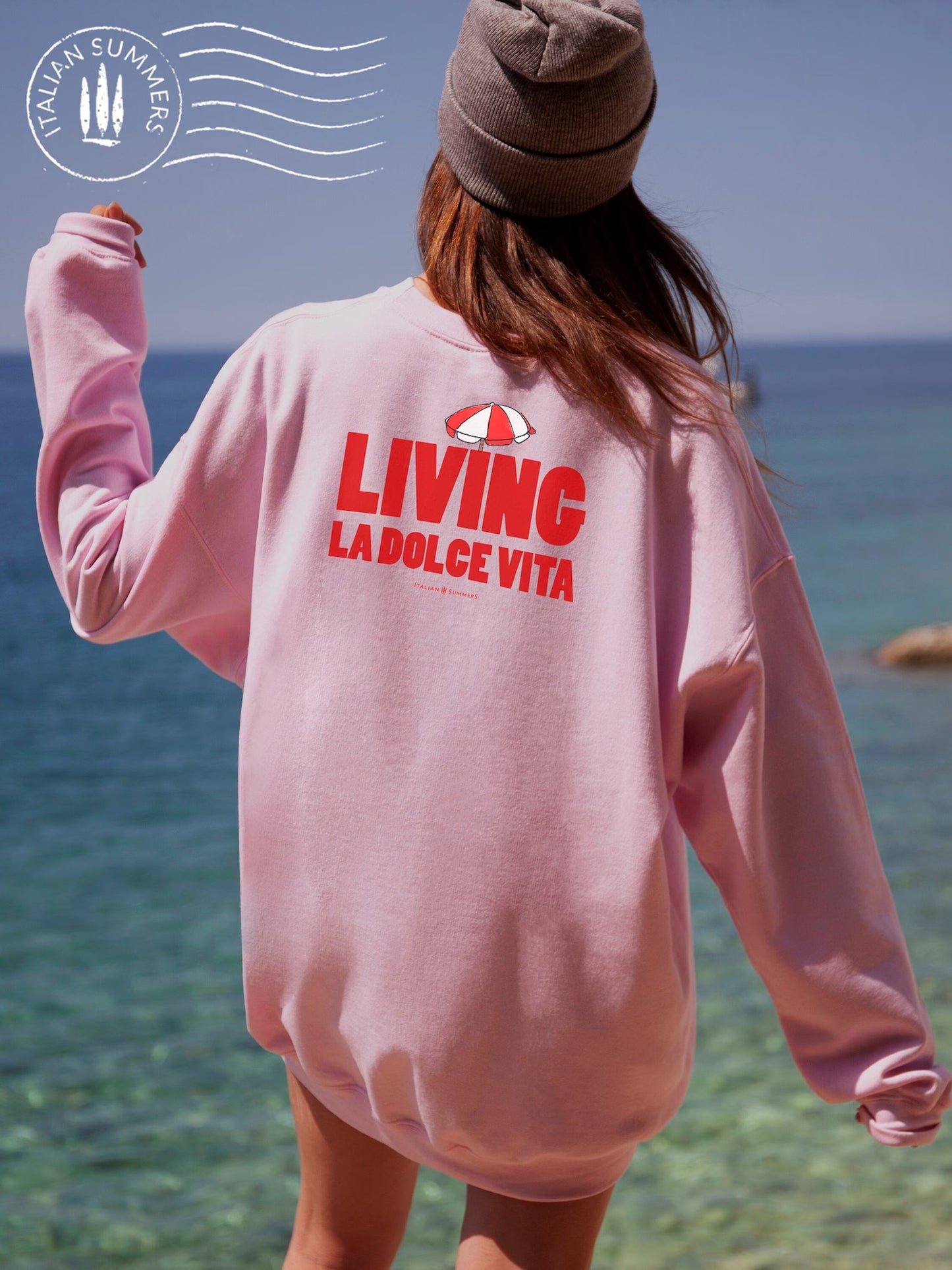  Soft and comfortable Italy-inspired  pink sweatshirt is embroidered in the front with the text 'Living la dolce Vita' in red bold letters and a small red and white beach umbrella on top. It features a large full-size print in the back with the same text and the same red and white beach umbrella. 