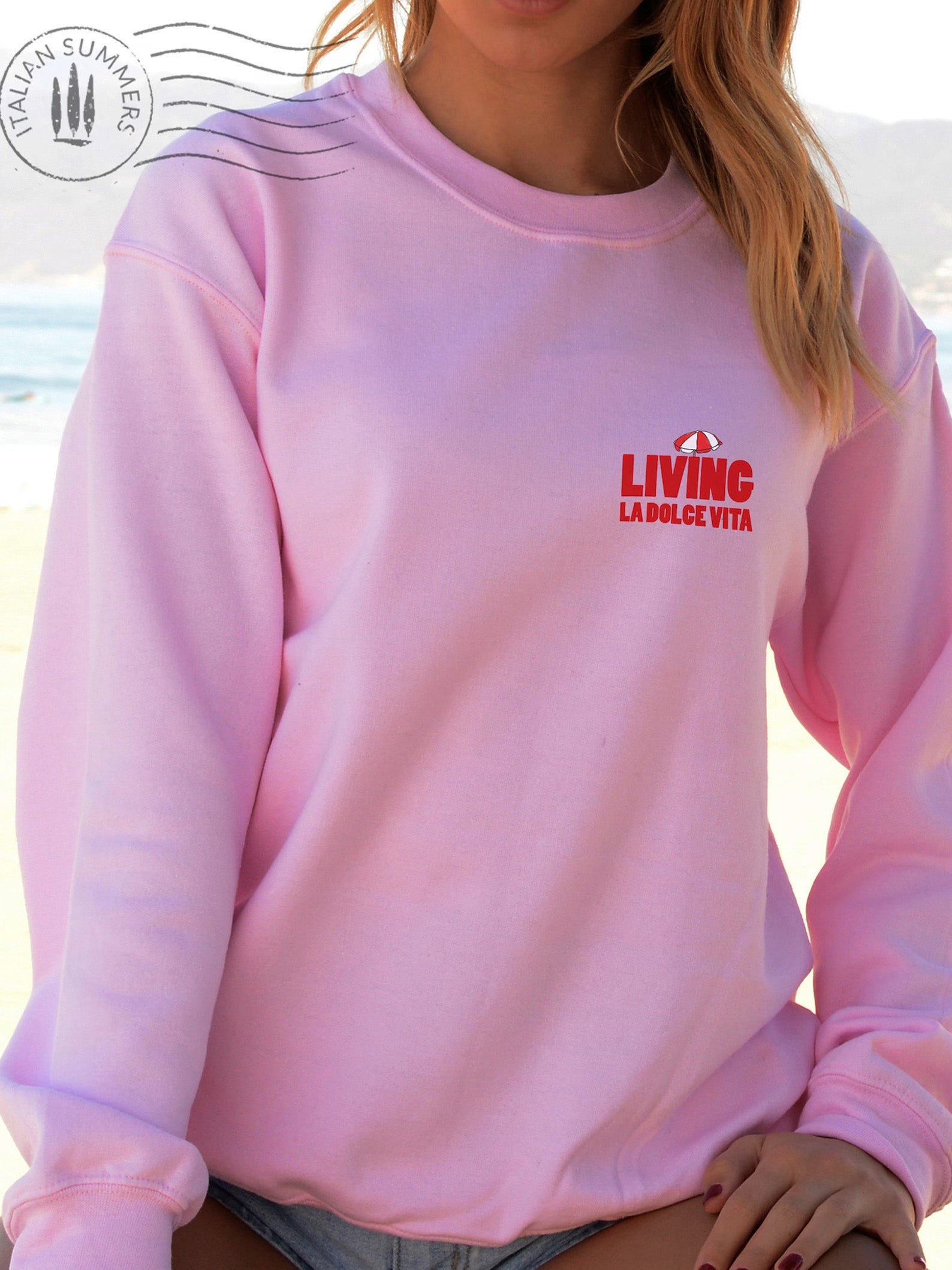  Soft and comfortable Italy-inspired  pink sweatshirt is embroidered in the front with the text 'Living la dolce Vita' in red bold letters and a small red and white beach umbrella on top. It features a large full-size print in the back with the same text and the same red and white beach umbrella. 