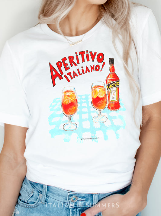 Italy inspired white unisex cotton T shirt with a print of a watercolor illustration of a bottle of red Italian aperritif and two spritz glasses filled with  ice, orange slices and spritz. A large red hand-painted text states: " Aperitivo Italiano" 
