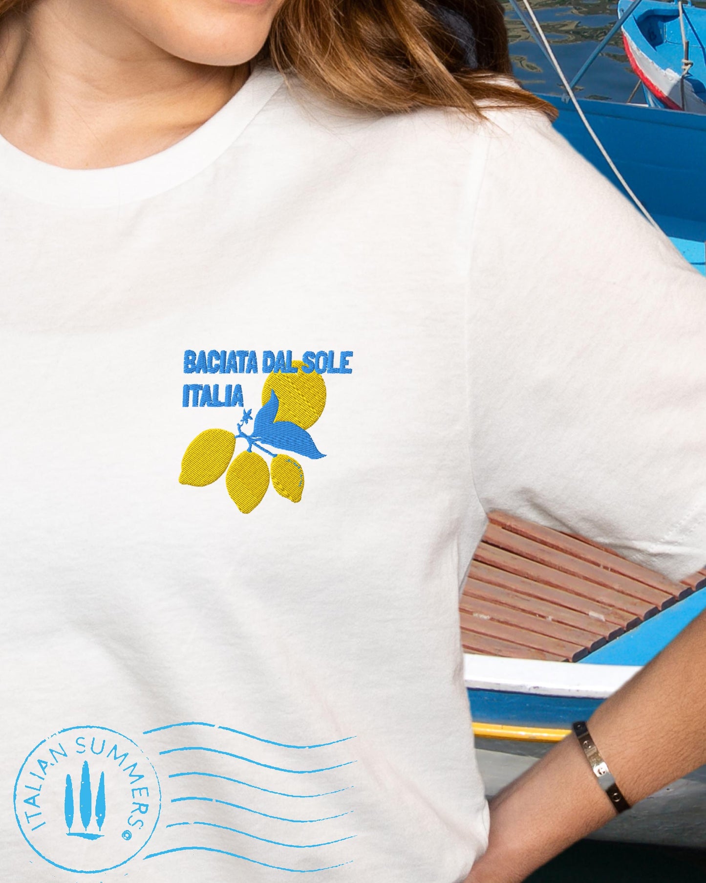 A bright graphic composition printed on a cotton shirt, featuring the Italian text: BACIATADALSOLE, 'kissed by the sun'. The aqua blue text is accompanied by three stylized Sorrento lemons with leaves and a flower against a yellow Sun.