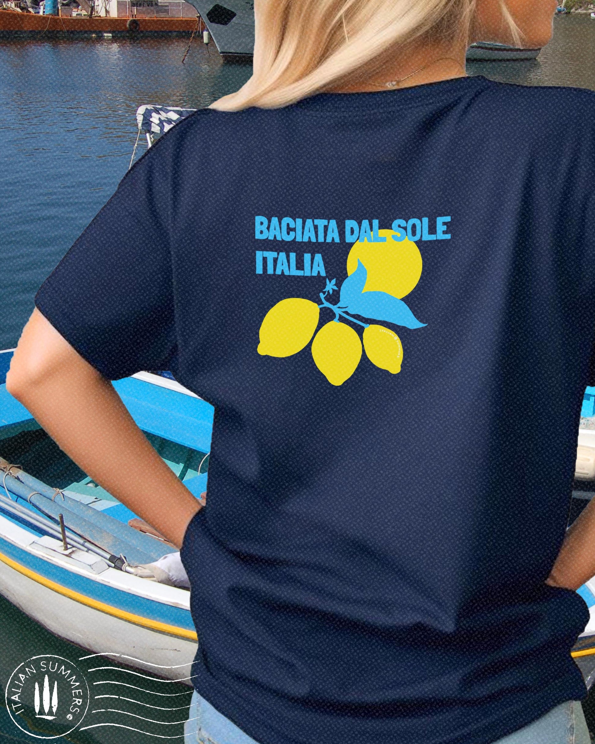 A bright graphic composition printed on a cotton shirt, featuring the Italian text: BACIATADALSOLE, 'kissed by the sun'. The aqua blue text is accompanied by three stylized Sorrento lemons with leaves and a flower against a yellow Sun.