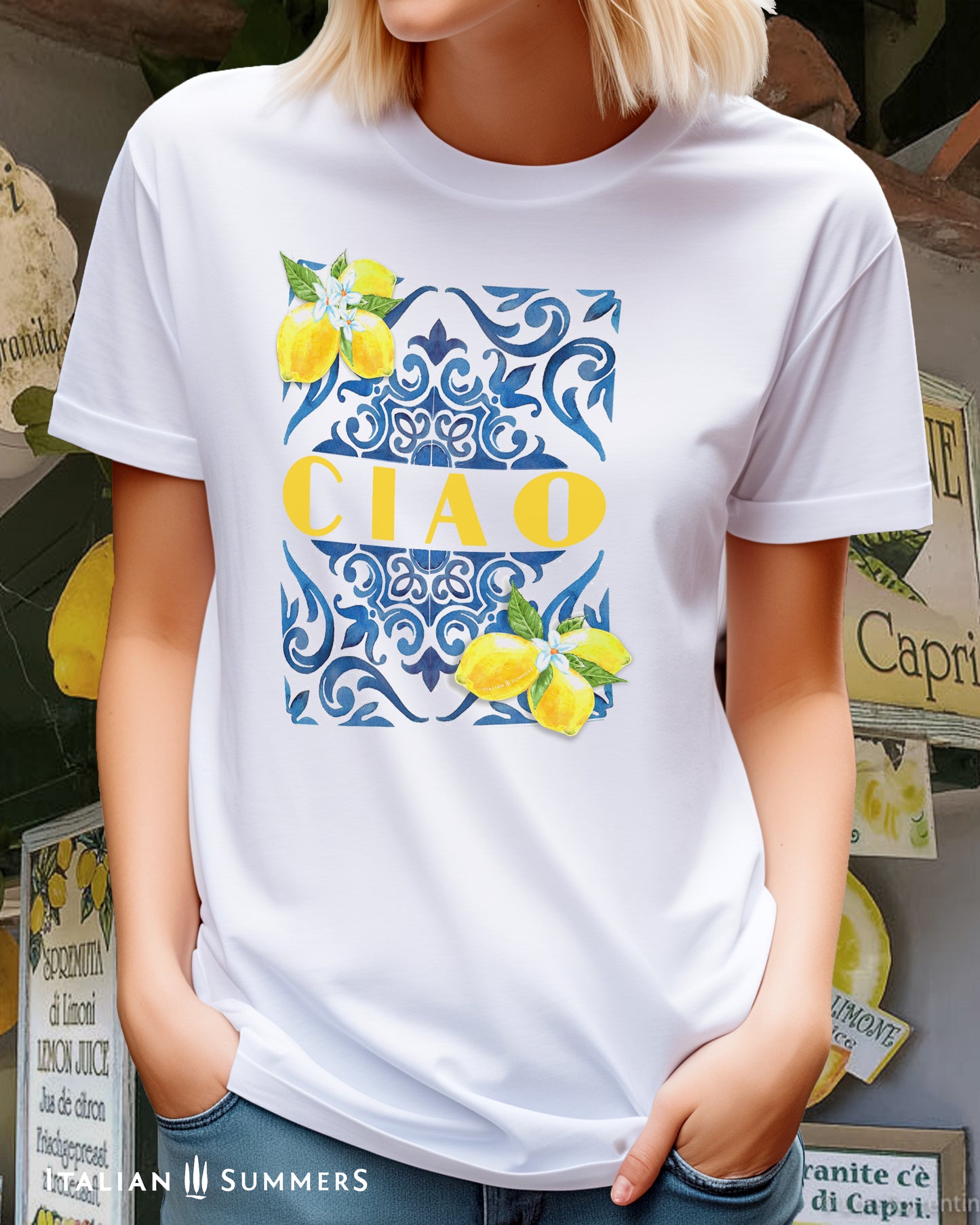 A blonde girl wears a white cotton T shirt with the print of a blue Italian Maiolica tile on the front, framed by lemons with blooms and the word CIAO  in large Art Deco yellow characters in the middle of the composition