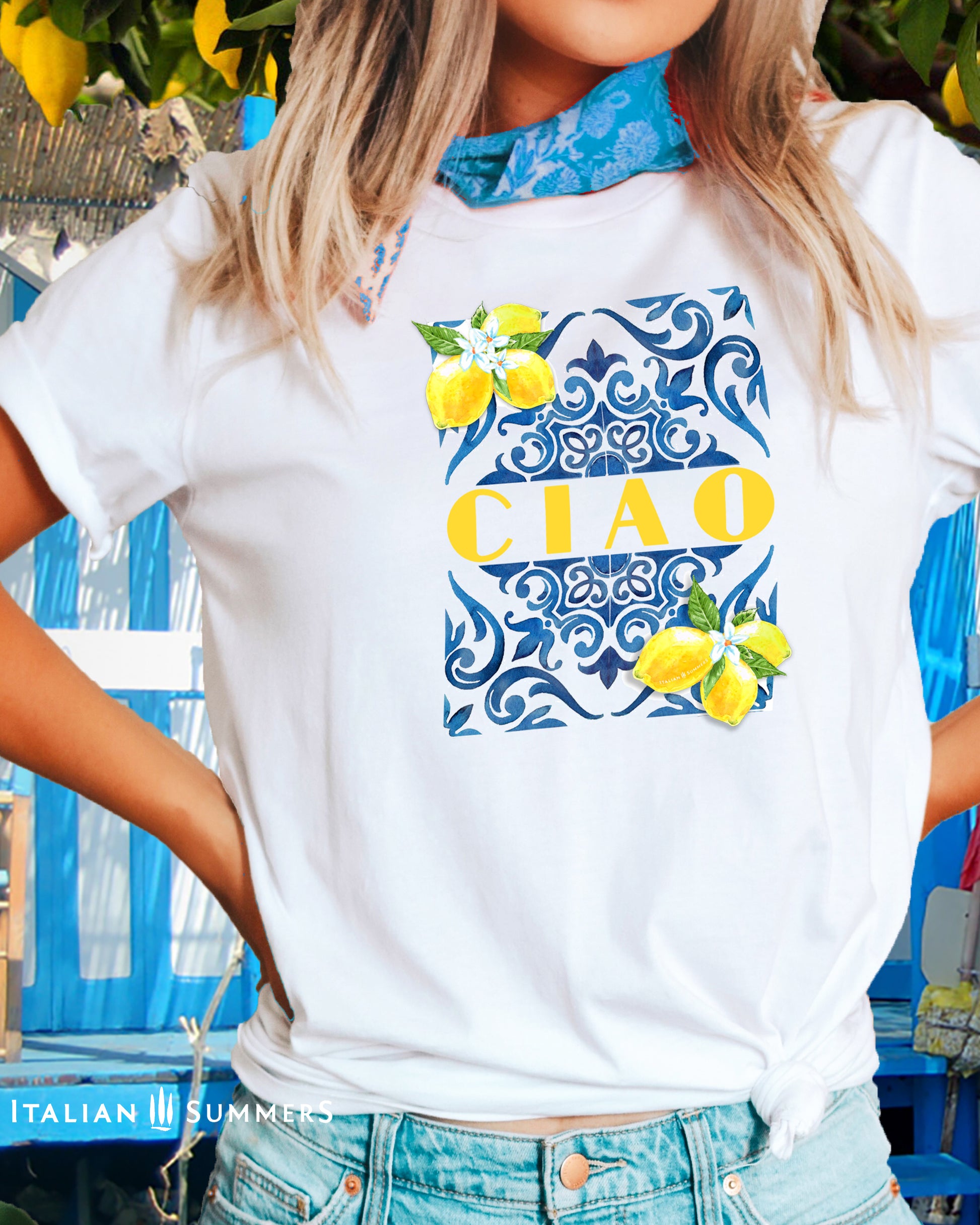 A blonde girl wears a white cotton T shirt with the print of a blue Italian Maiolica tile on the front, framed by lemons with blooms and the word CIAO  in large Art Deco yellow characters in the middle of the composition