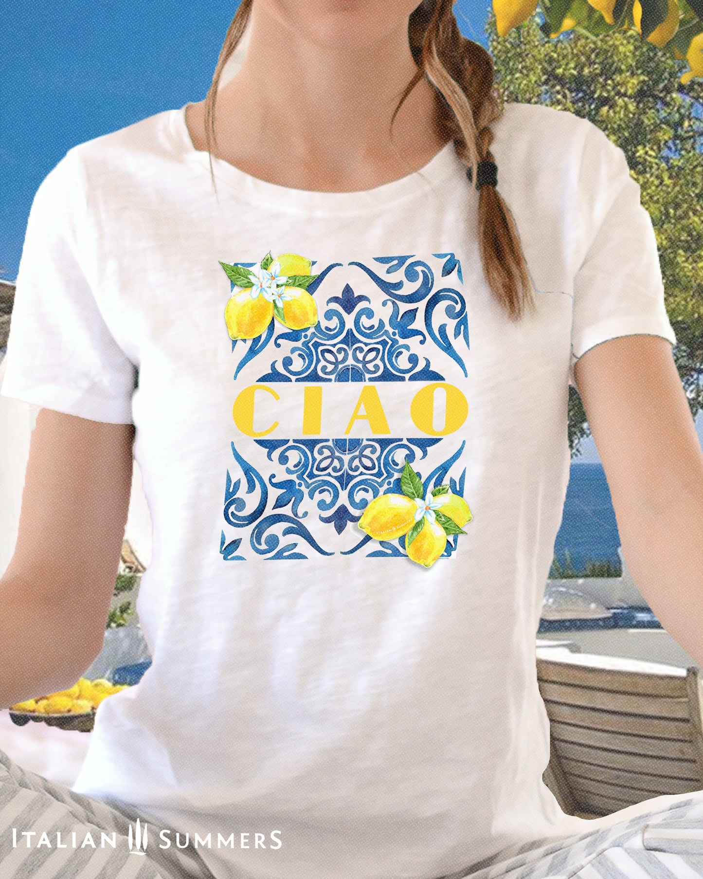 A dark blonde  girl wears a white cotton T shirt with the print of a blue Italian Maiolica tile on the front, framed by lemons with blooms and the word CIAO  in large Art Deco yellow characters in the middle of the composition
