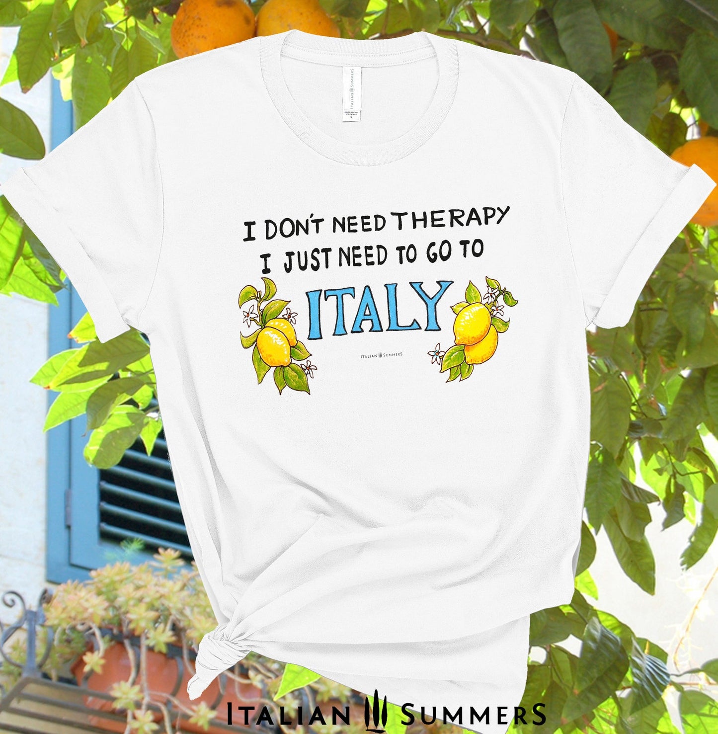 Italy inspired white cotton T shirt with the quote " I don't need therapy I just need to go to Italy" the word " Italy" Is in capital blue letters and flanked by hand-painted bunches of lemons with flowers. A bestseller made by Italian Summers