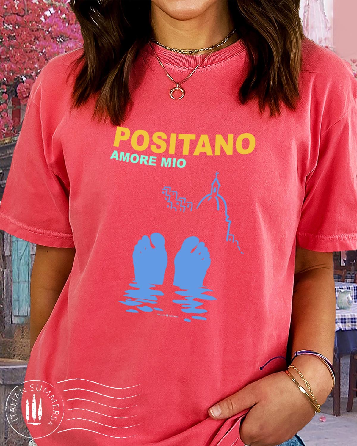 Italy-inspired garment-dyed  Coral colored T shirt with the text: POSITANO Amore Mio (Positano, my love) with a stylized view of the seaside town and the silhouette of two feet sticking out of the waters of the Mediterranean Sea. Made by Italian Summers