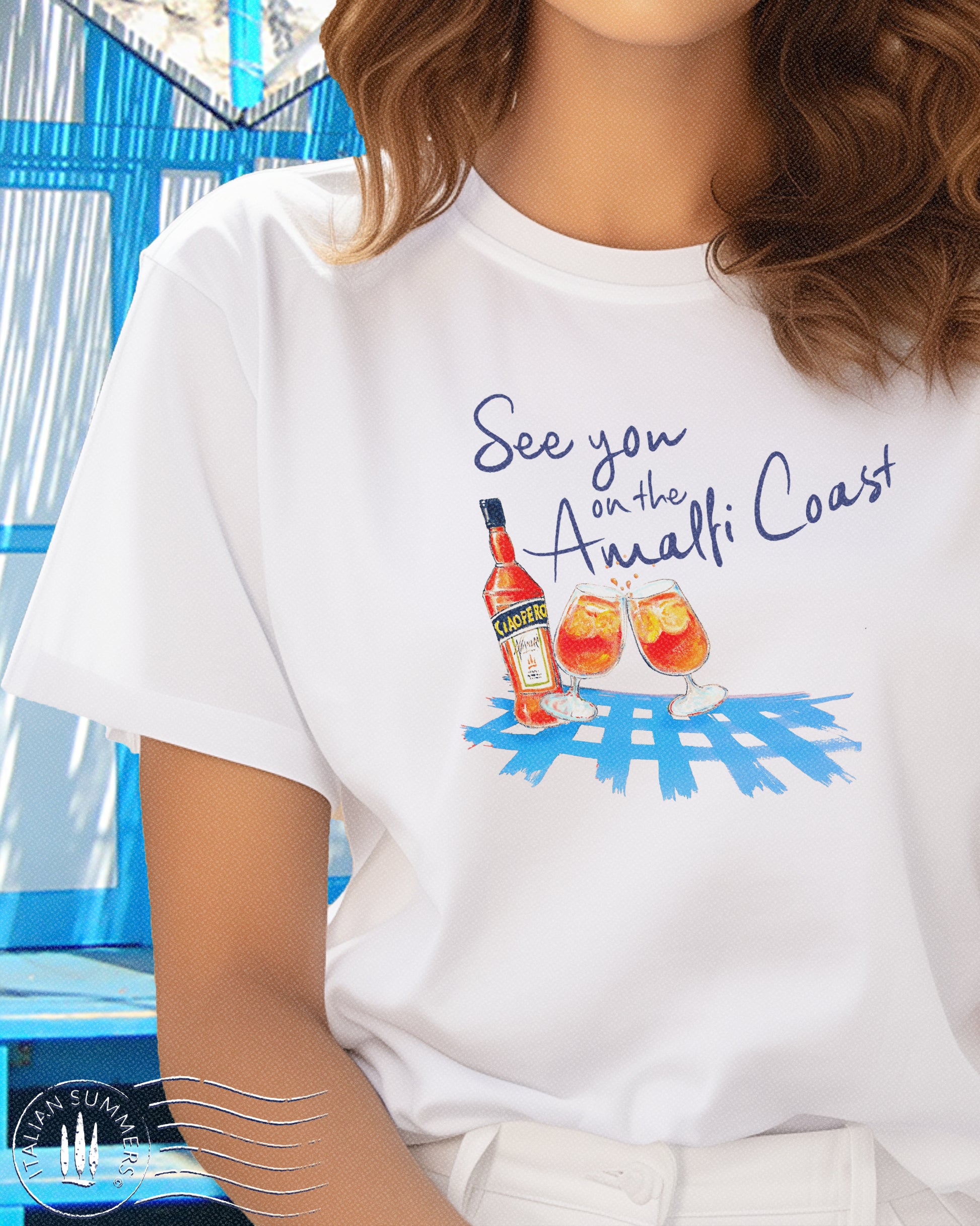 Italy inspired white t-shirt with the text See you on the Amalfi Coast in navy blue and a sketch in water color with an Aperol bottle and 2 glassis filled with Aperol Spritz on a bright blue table cloth.