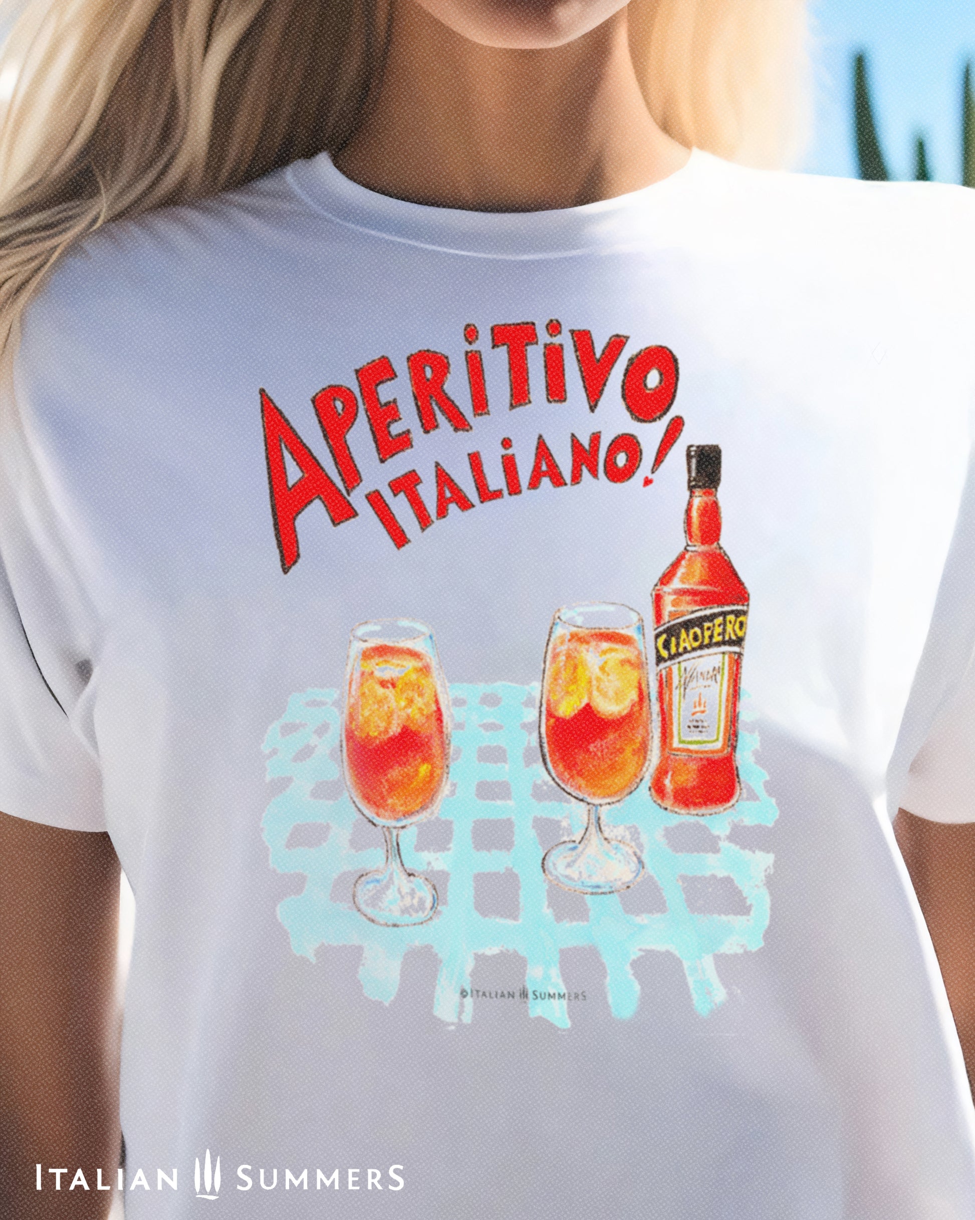 A woman wearing a white unisex cotton T shirt with a print of a watercolor illustration of a bottle of red Italian aperitif and two spritz glasses filled with  ice, orange slices and spritz. A large red hand-painted text states: " Aperitivo Italiano" 