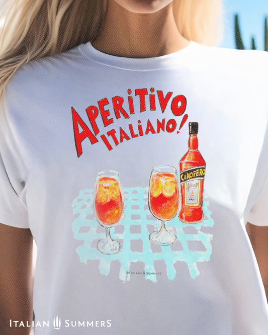 A woman wearing a white unisex cotton T shirt with a print of a watercolor illustration of a bottle of red Italian aperitif and two spritz glasses filled with  ice, orange slices and spritz. A large red hand-painted text states: " Aperitivo Italiano" 