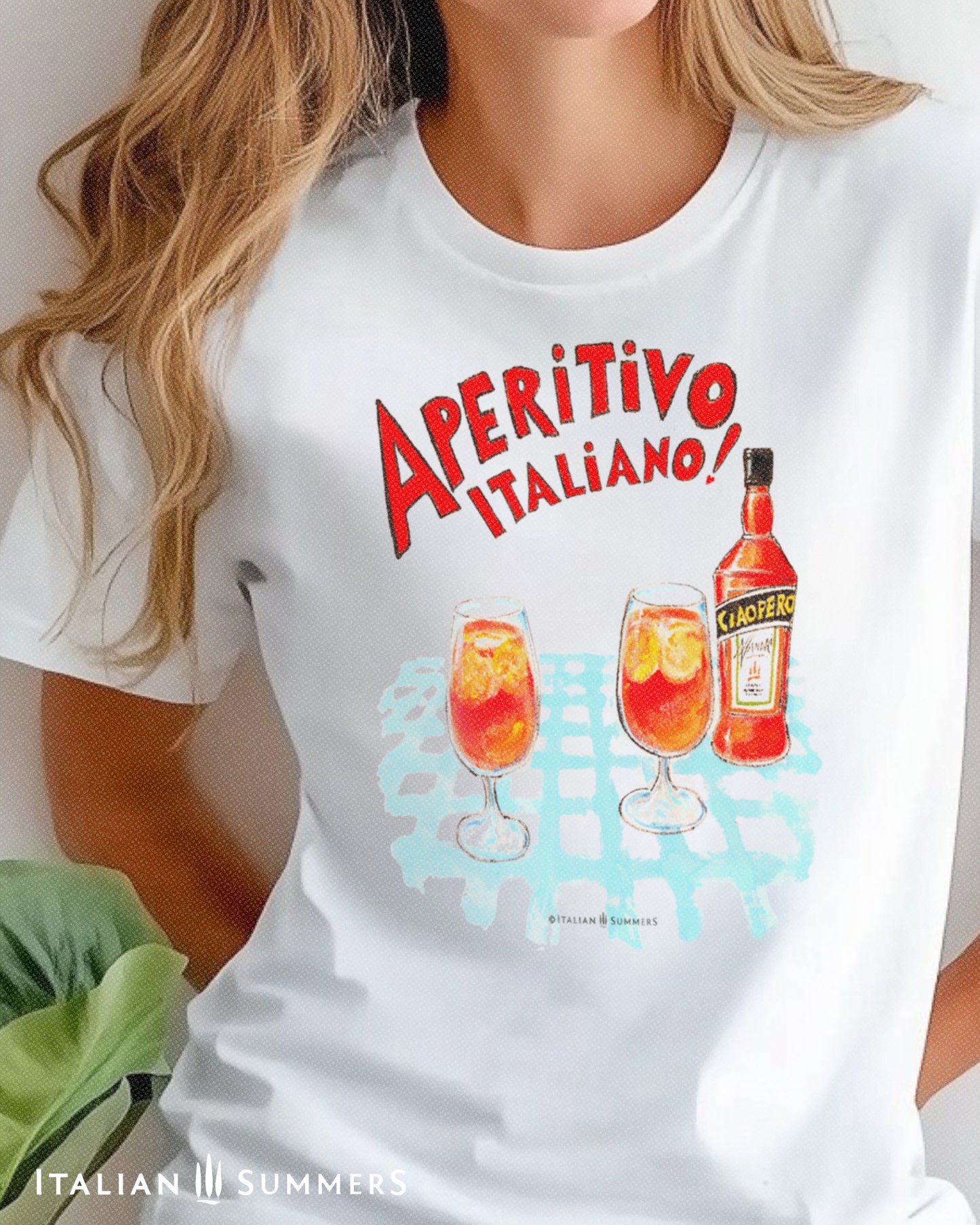 A woman wearing a white unisex cotton T shirt with a print of a watercolor illustration of a bottle of red Italian aperitif and two spritz glasses filled with  ice, orange slices and spritz. A large red hand-painted text states: " Aperitivo Italiano" 
