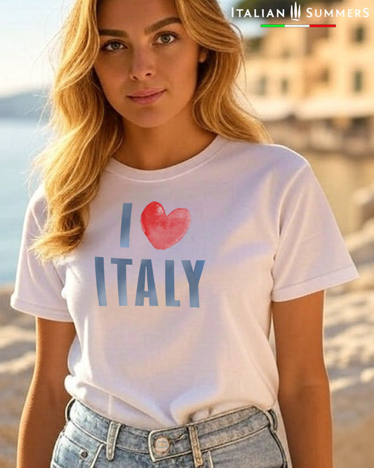 A woman by an Italian seaside location wears a white t shirt with the text I LOVE ITALY, the word "love" is a red watercolor heart shape