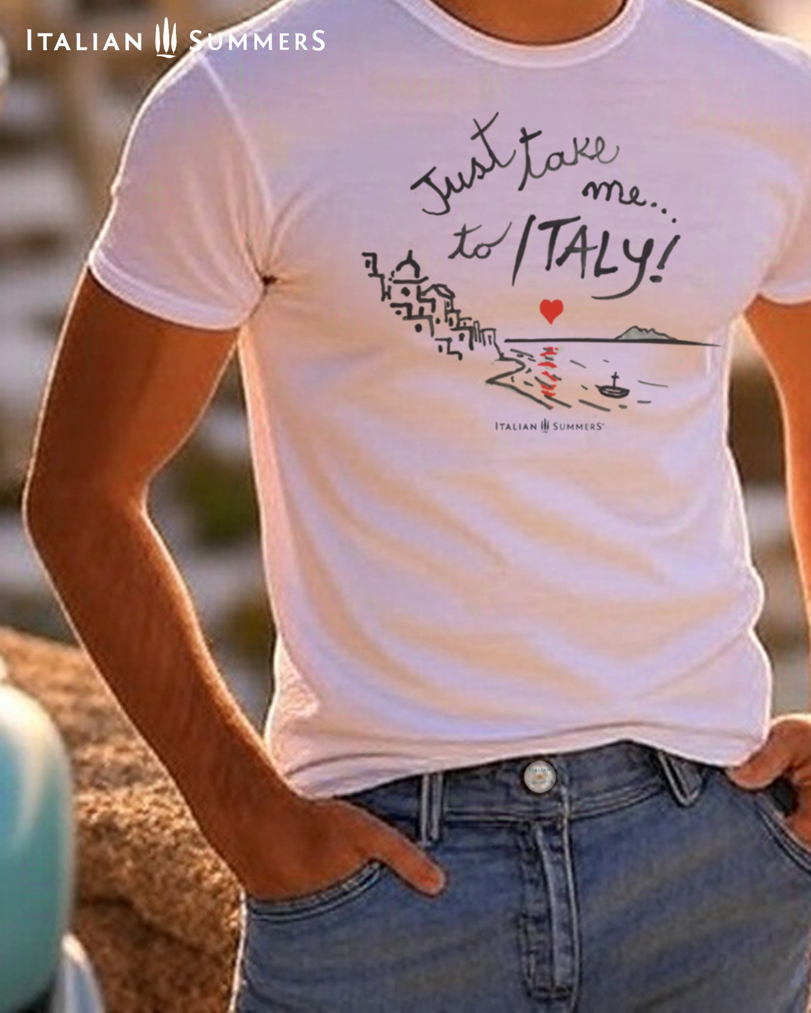 An Italian young man  wearing a Italy-inspired Unisex crew neck T shirt with a print of a hand-painted  stylized Amalfi Coast village with a red heart setting on the sea and reflecting its glow on the waters. The hand-painted phrase "Just take me to Italy" is above the composition 