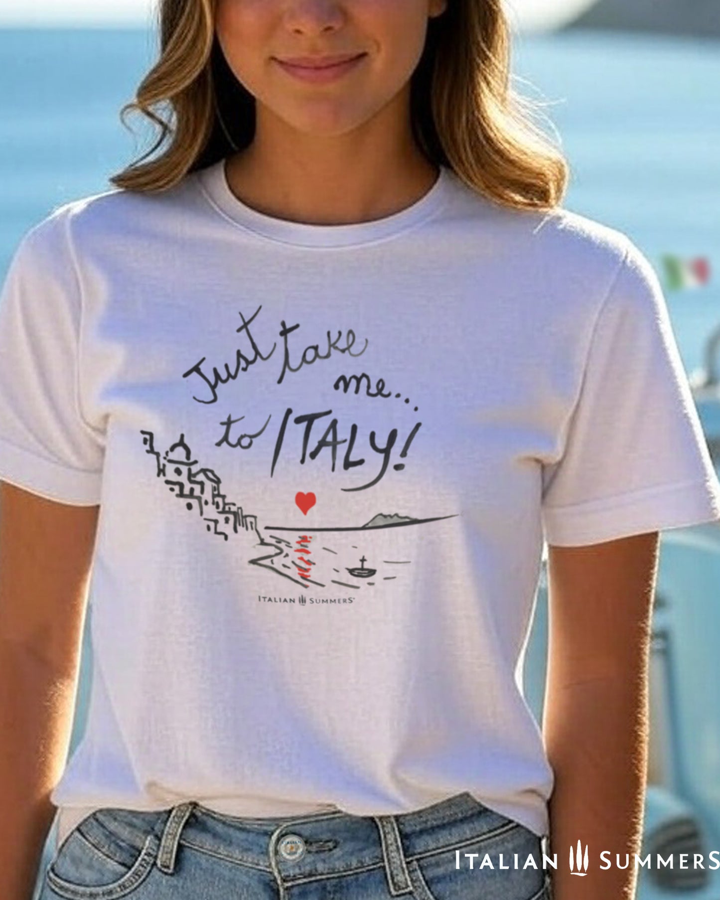 An Italian young woman  wearing a Italy-inspired Unisex crew neck T shirt with a print of a hand-painted  stylized Amalfi Coast village with a red heart setting on the sea and reflecting its glow on the waters. The hand-painted phrase "Just take me to Italy" is above the composition 