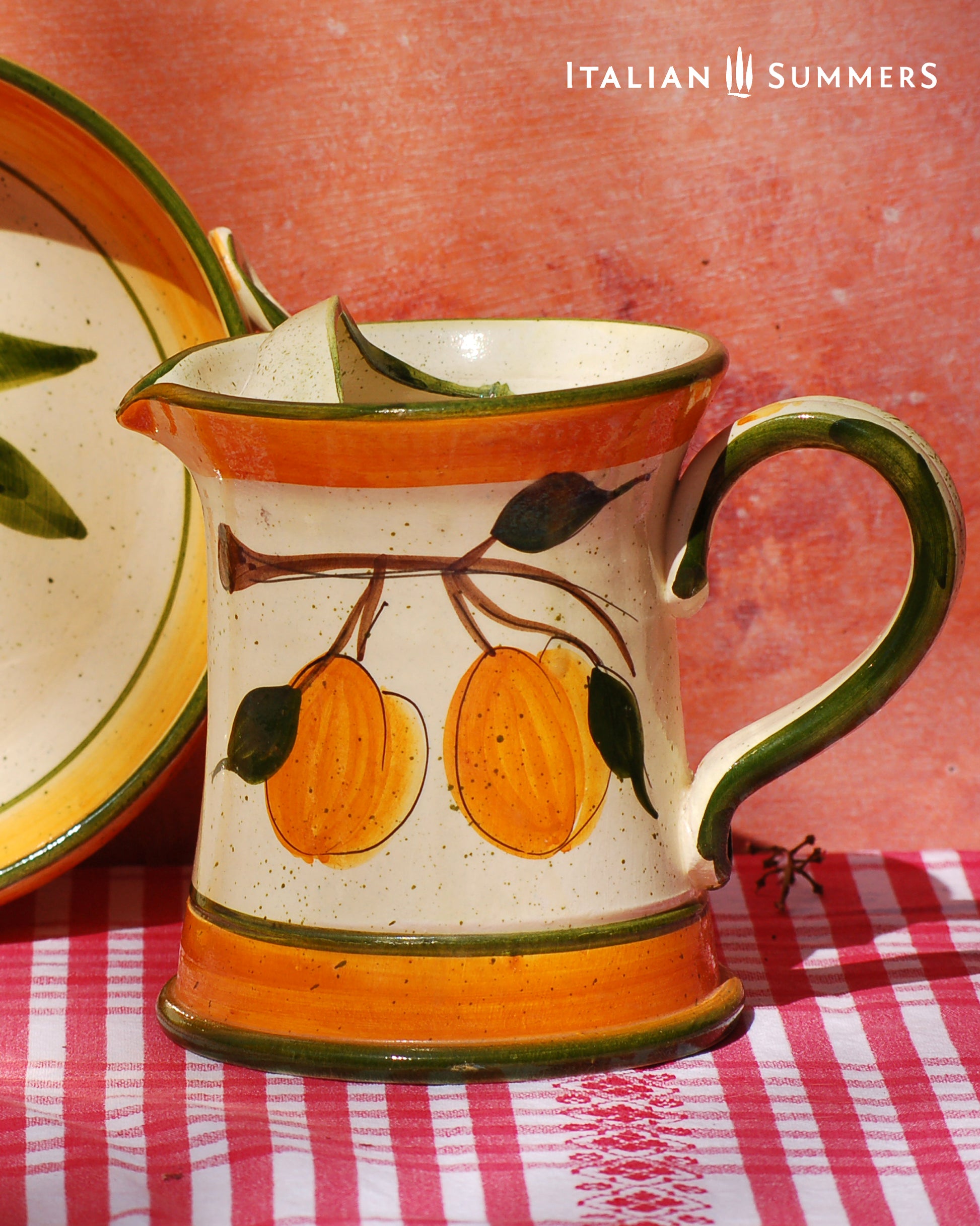 Ceramic pitcher with fruit design on a checkered tablecloth, featuring 'Italian Summers' branding.
