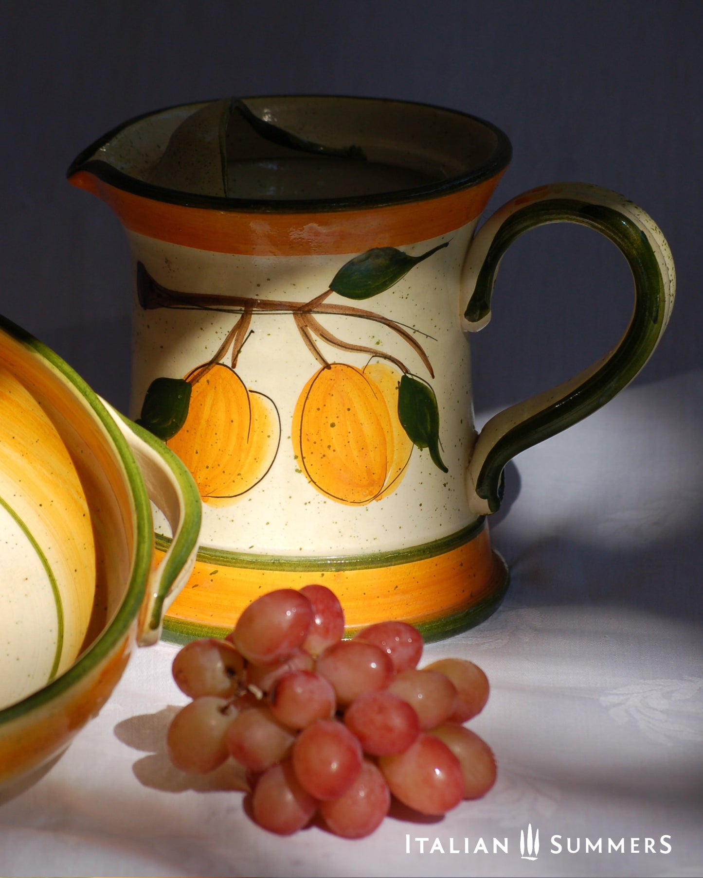 Ceramic pitcher with fruit design on a dark background, accompanied by grapes and another ceramic item.
