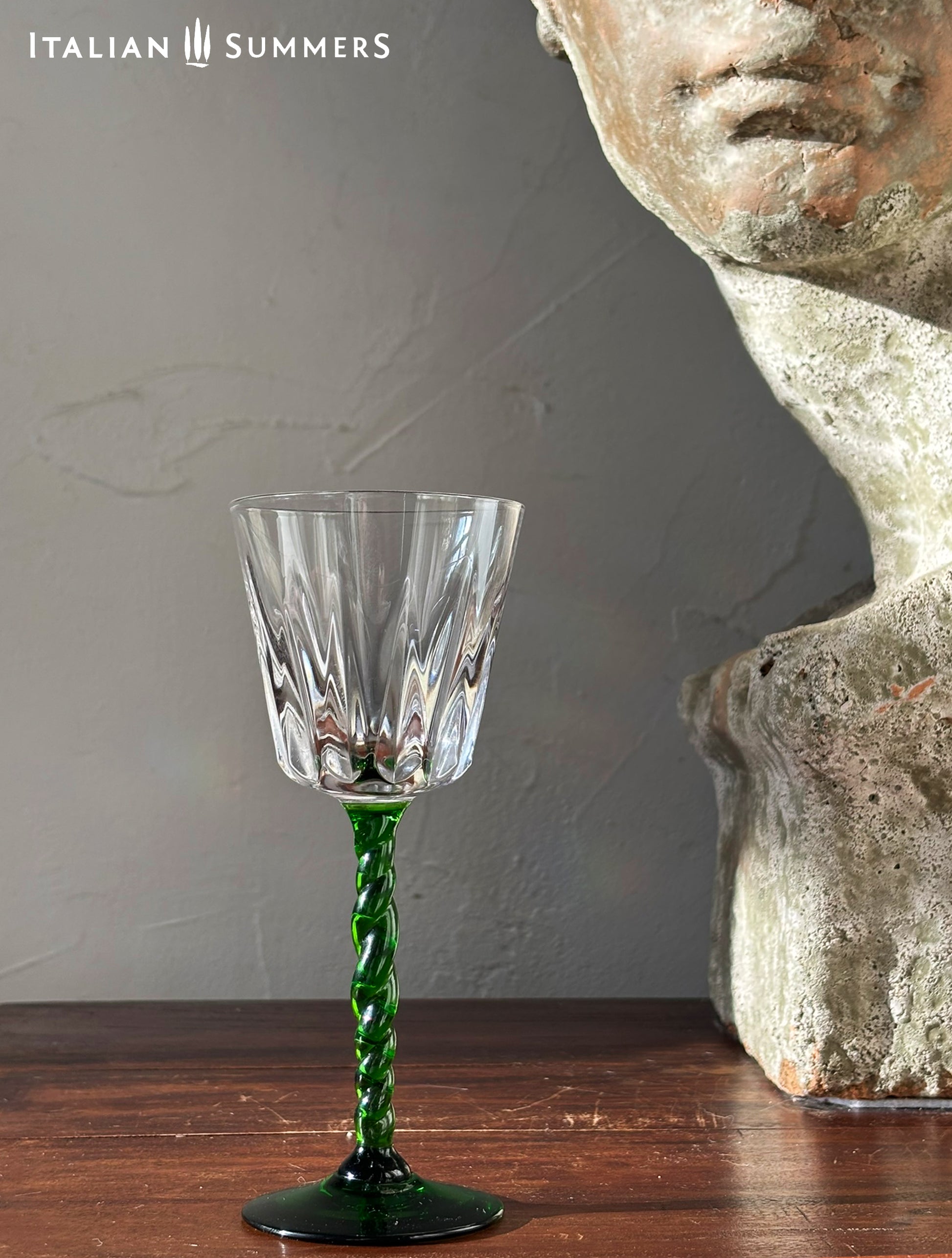 Crystal wine glass with green stem on a wooden surface next to a stone sculpture, with 'Italian Summers' branding.